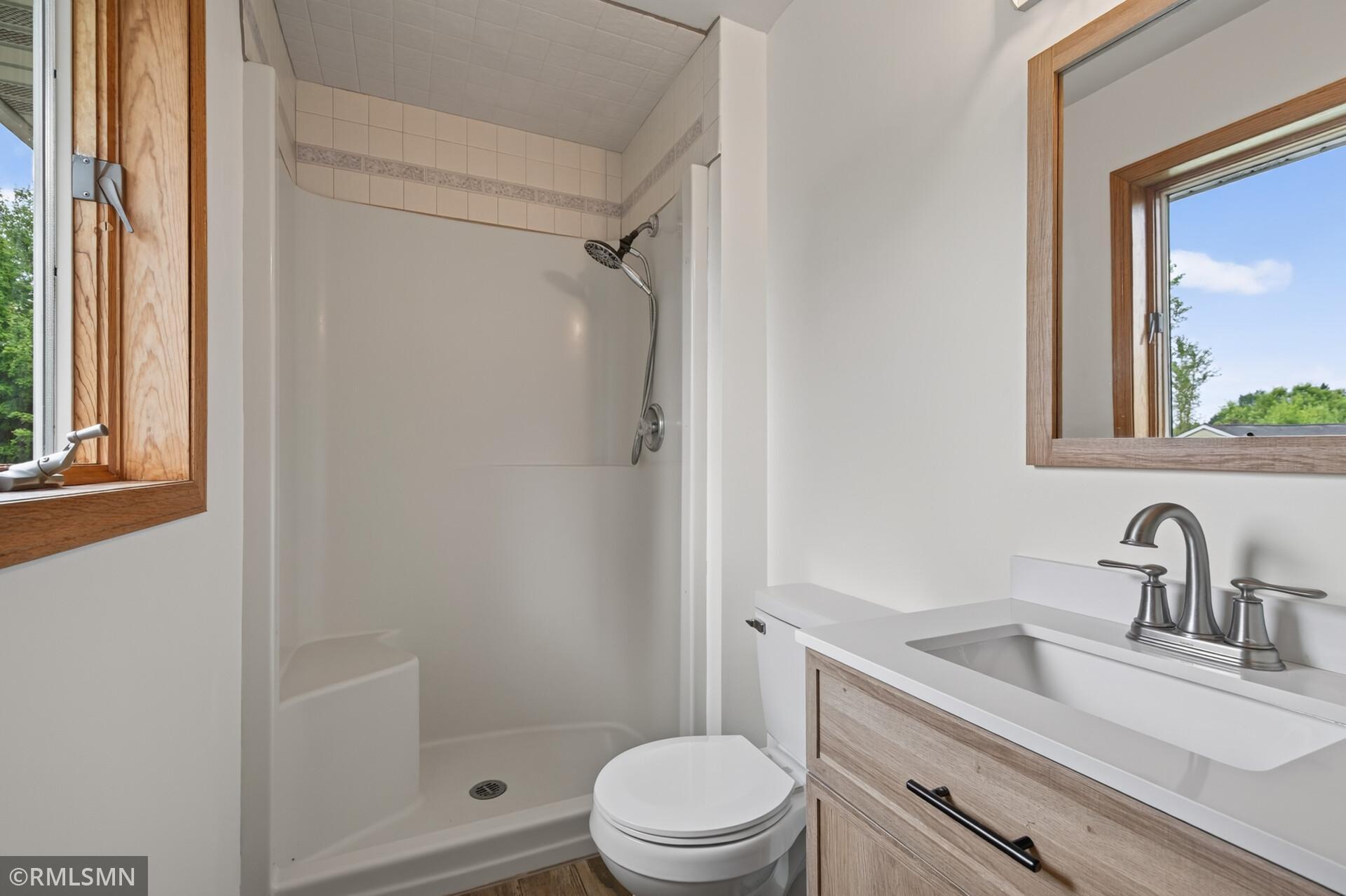 Bathroom with a white shower, toilet, and light wood vanity beside wood-trimmed windows.