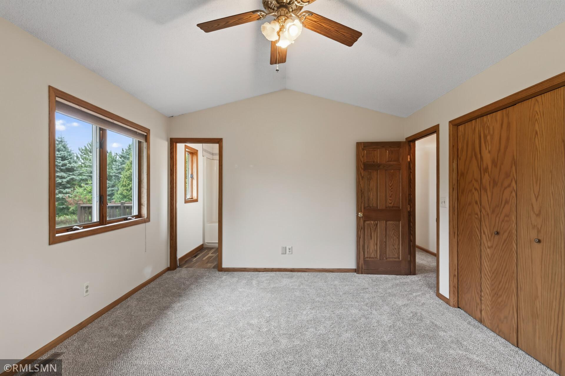 Empty bedroom with gray carpet, a ceiling fan, and wooden closet doors beside a window.