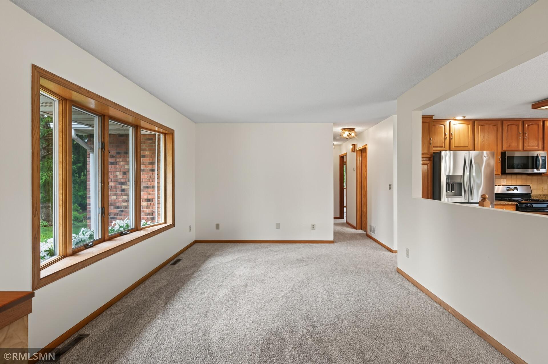 Empty carpeted living room with large wood-trimmed windows and an adjoining kitchen.