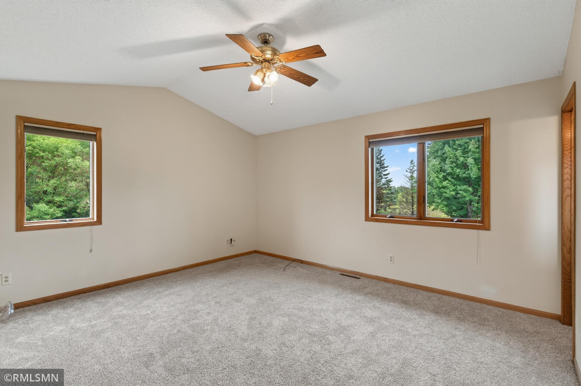 Empty carpeted bedroom with wood-trimmed windows overlooking trees and a ceiling fan.