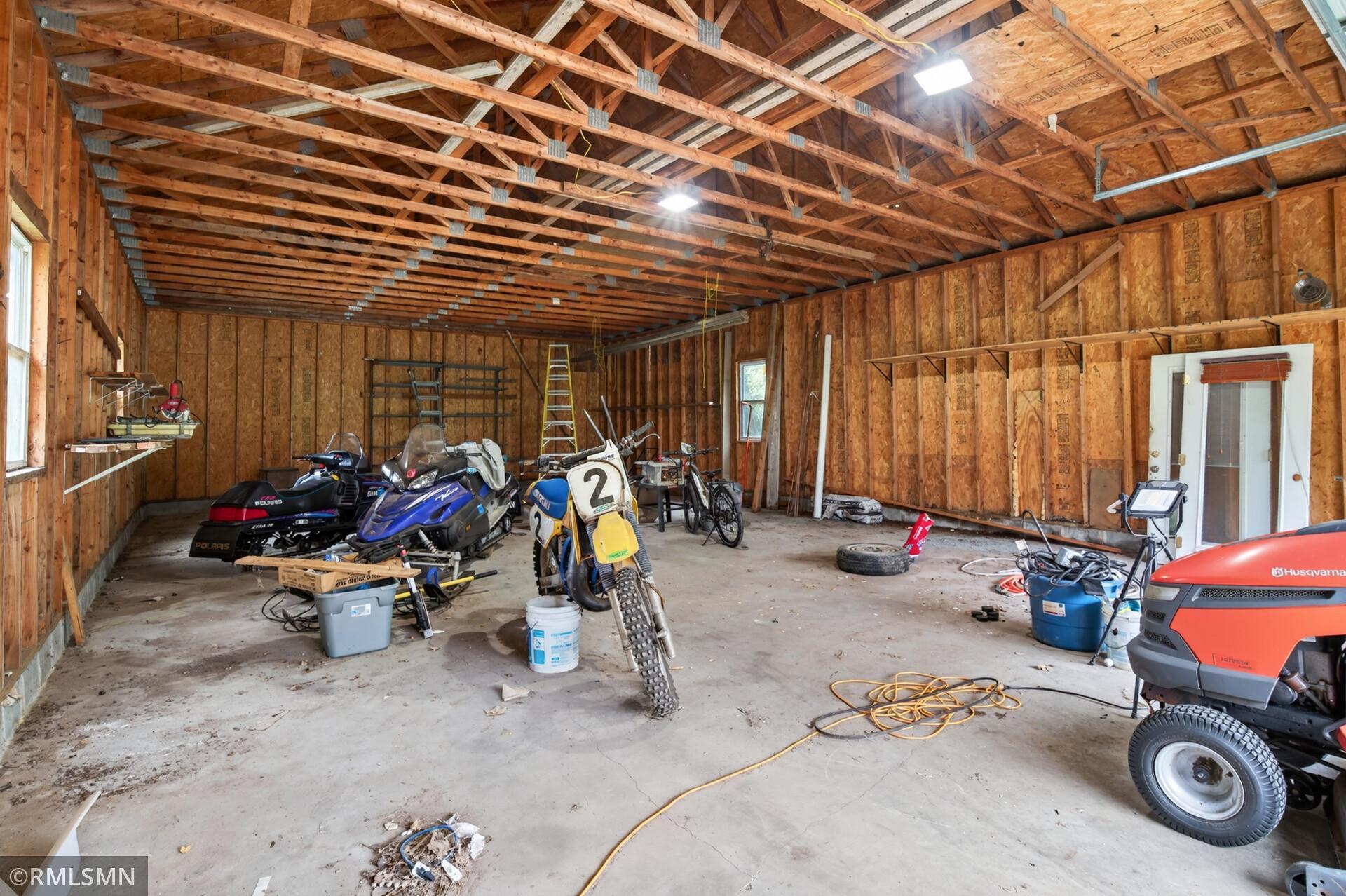 Large unfinished garage with exposed roof trusses, snowmobiles, motorcycles, and a riding mower.