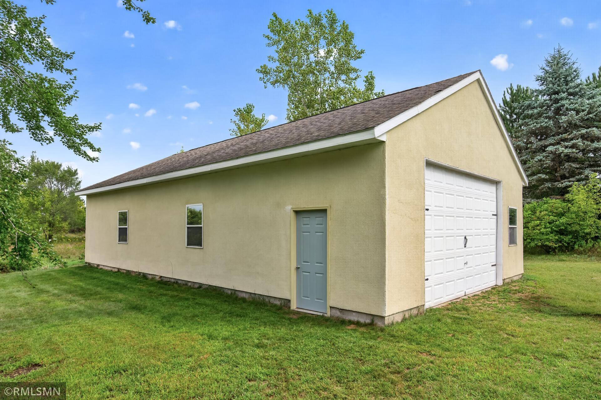 Beige detached garage with a white overhead door and blue-gray side entrance, surrounded by lawn and trees.