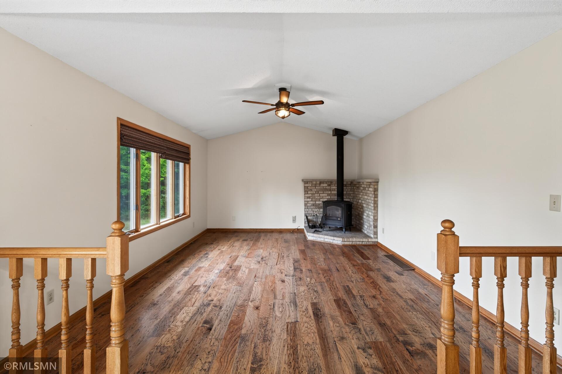 Vacant living area with hardwood floors, vaulted ceiling, and a wood-burning stove in a brick surround.