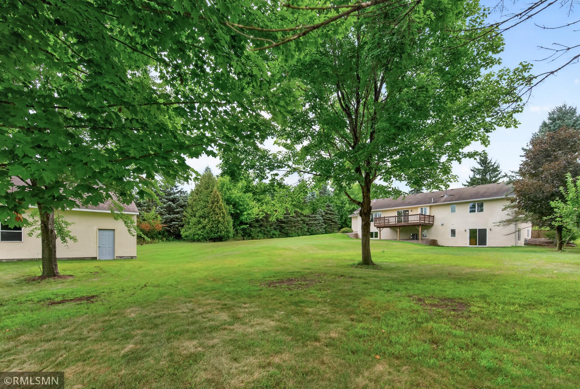 Rear of a beige home with a raised deck, detached outbuilding, and broad tree-shaded lawn.