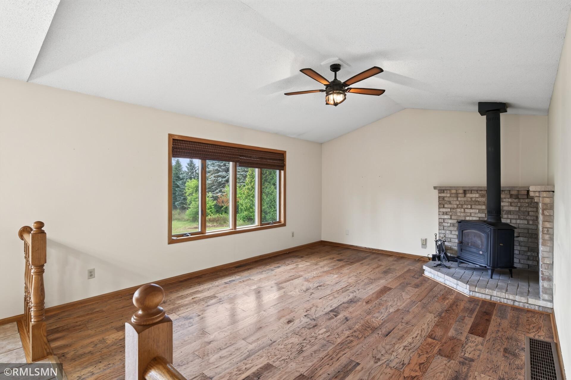 Empty living room with hardwood floors, a wood stove in a brick fireplace, and a large window overlooking trees.