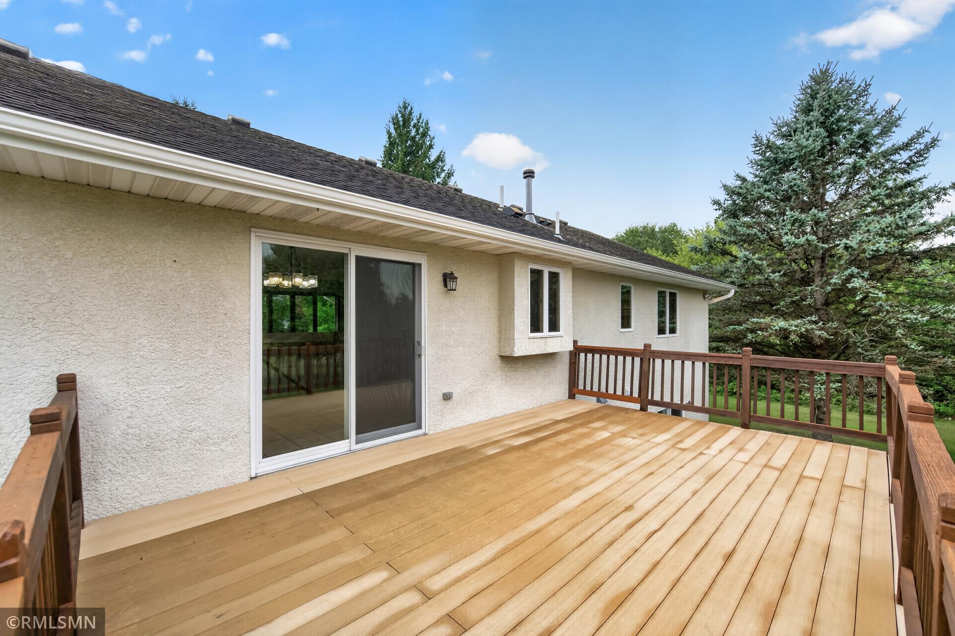 Wooden deck along a cream stucco house, with brown railings and evergreen trees beyond.