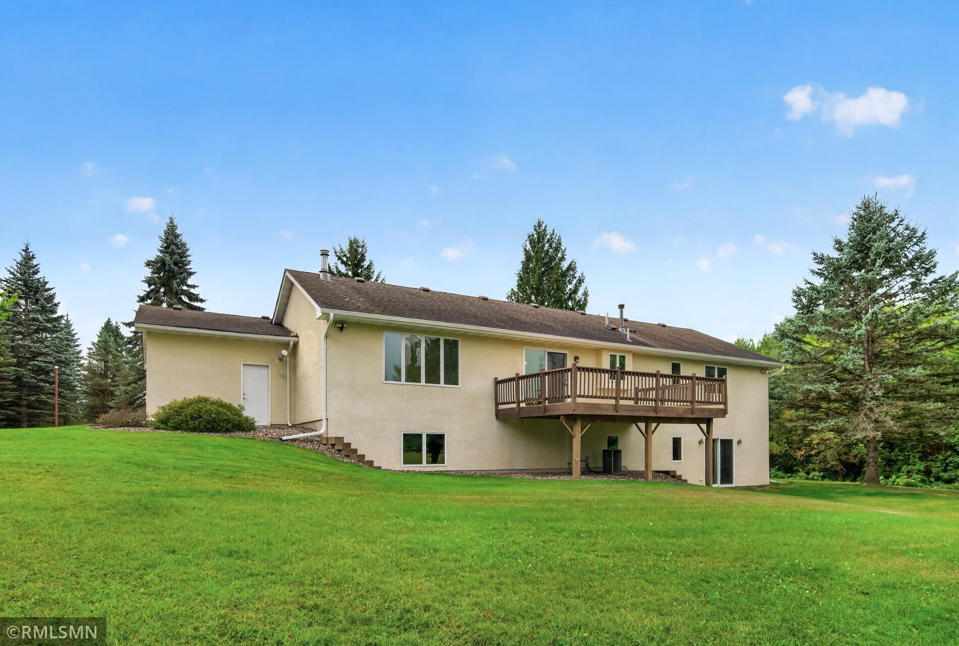 Beige house with a raised wooden deck overlooking a broad lawn and mature evergreen trees.