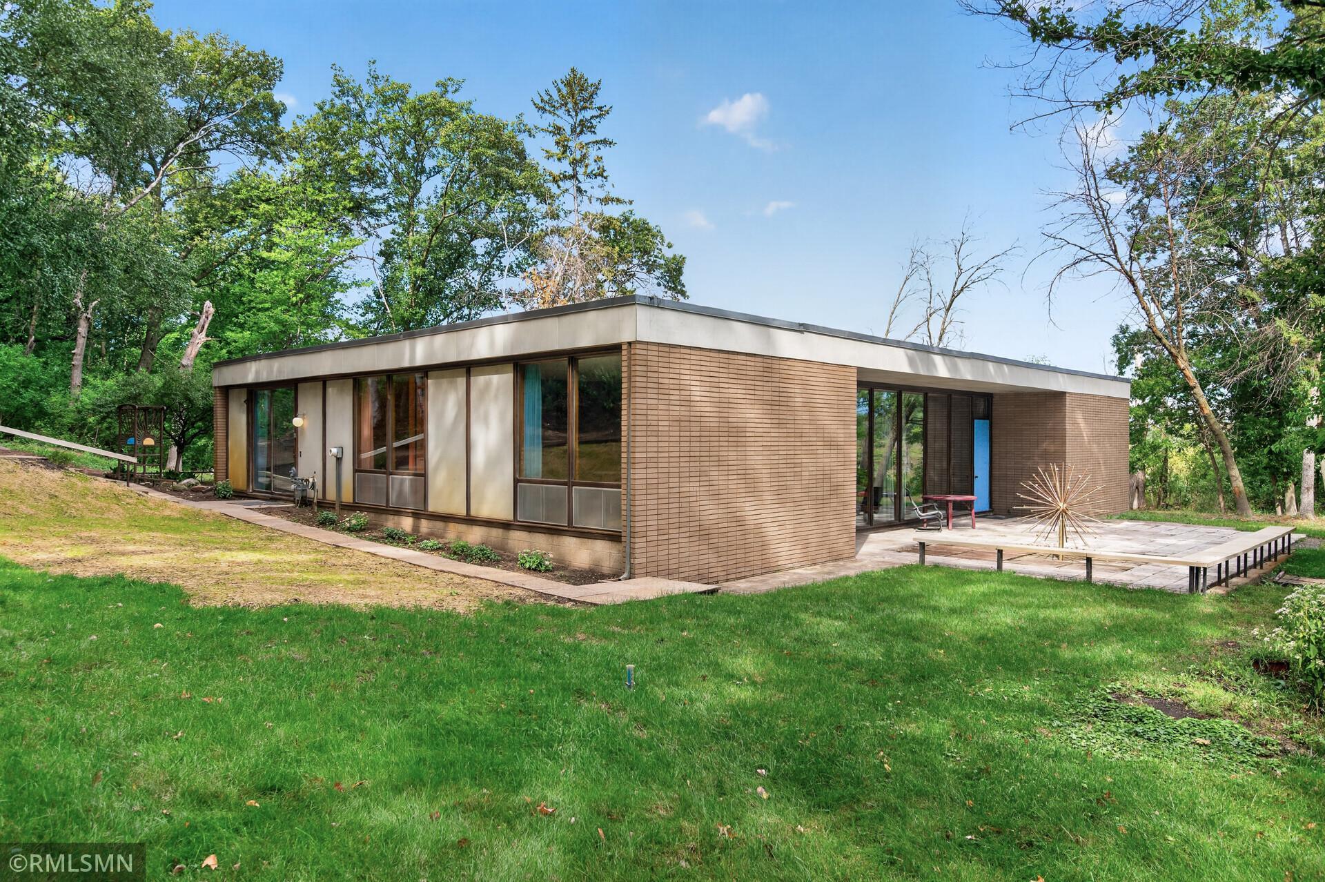 Flat-roofed modernist brick home with broad windows and a paved patio, surrounded by trees.