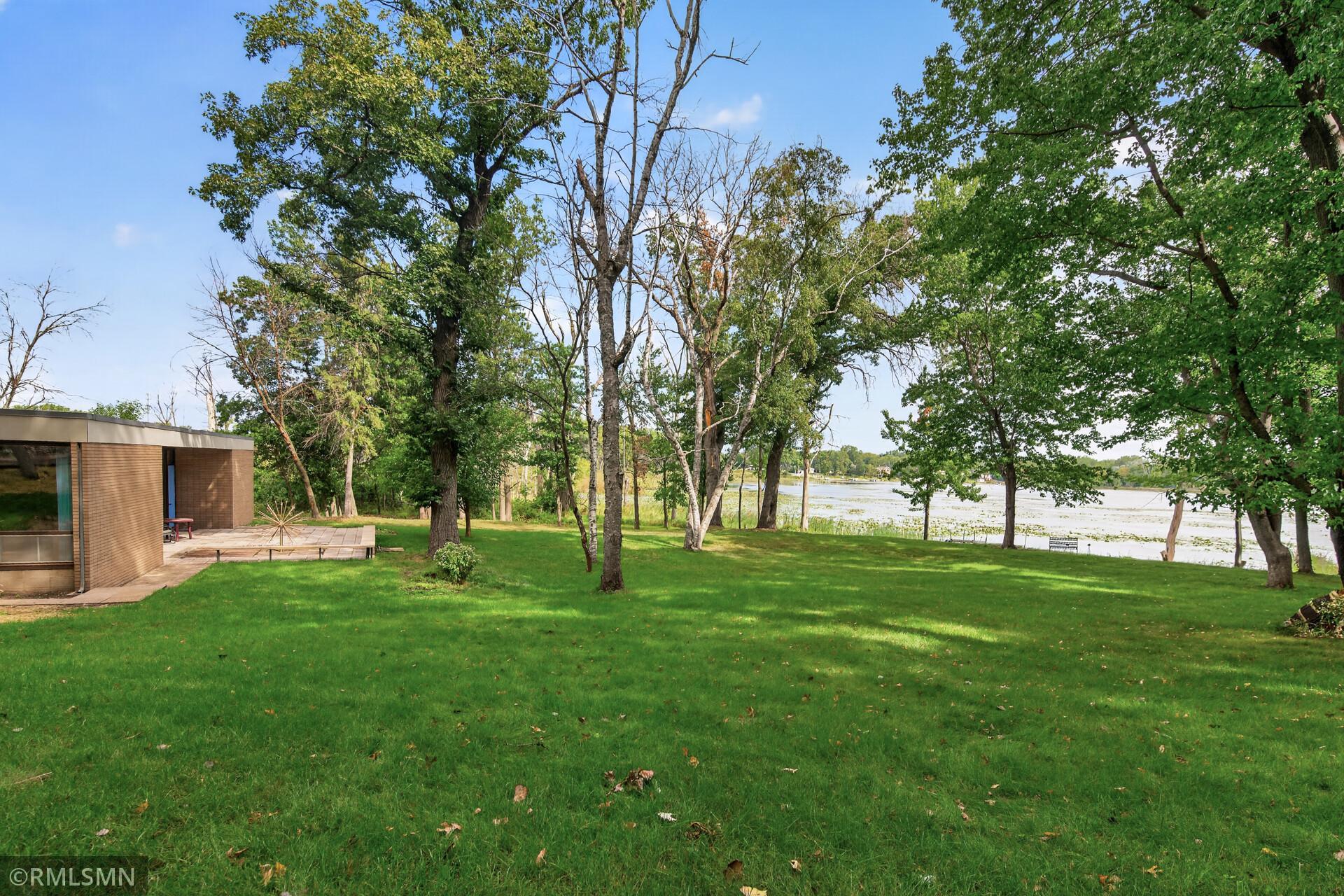 Tree-shaded lawn beside a lake, with a modern building and patio at the left.