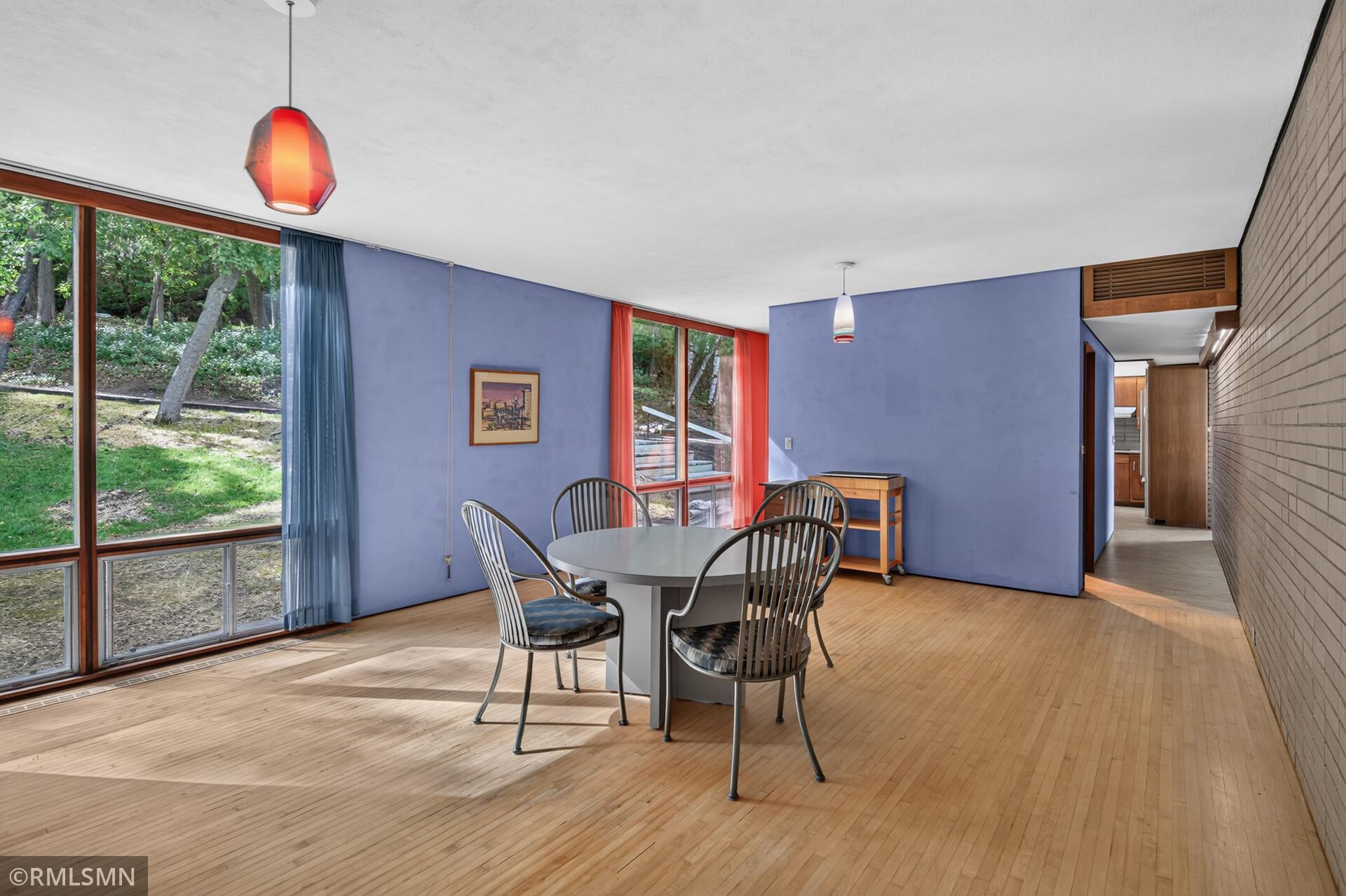Dining area with a round table and metal chairs, purple walls, and large windows overlooking trees.