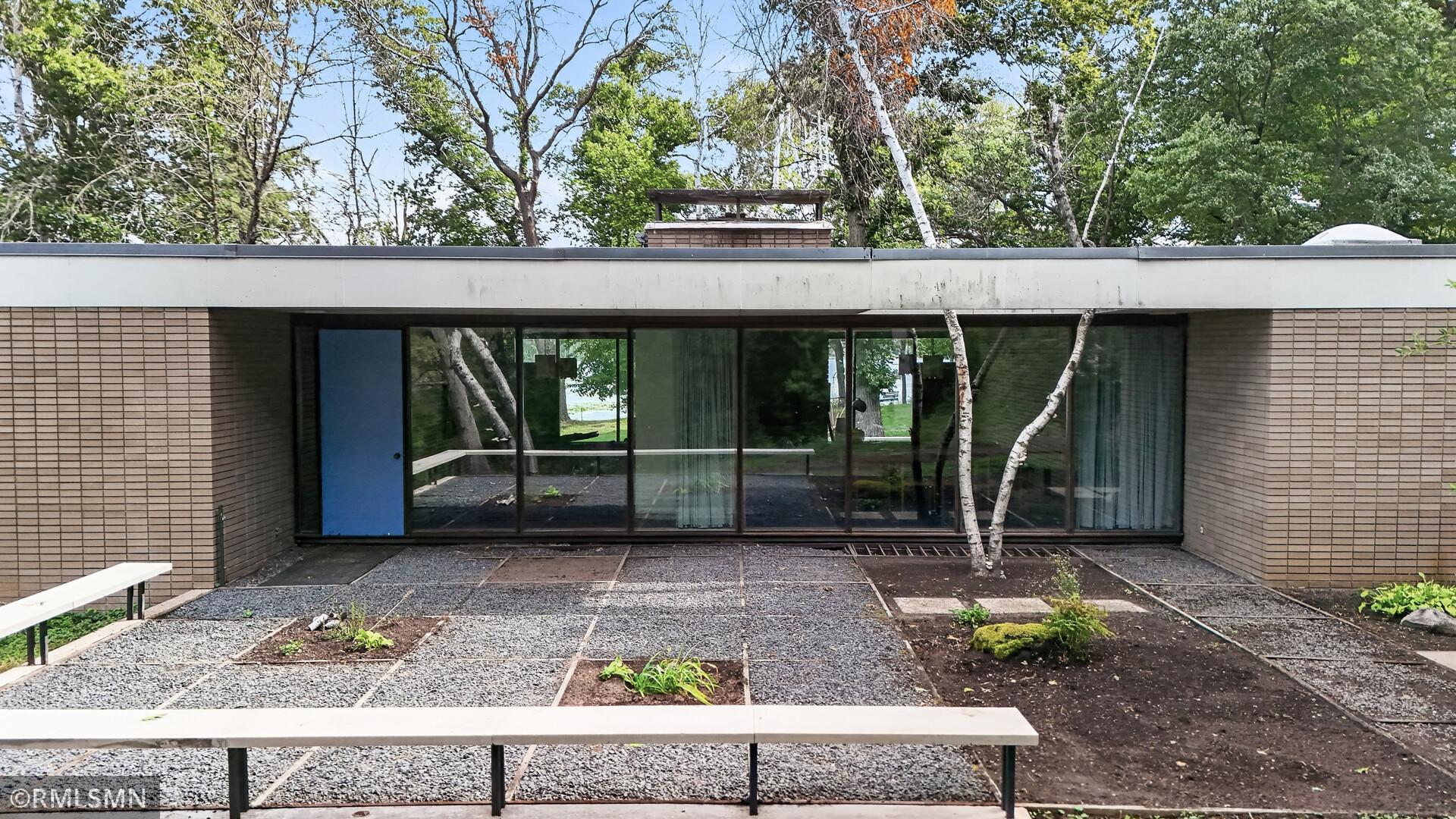 Modern brick home with a flat roof, expansive glass doors, and a gravel courtyard beneath mature trees.