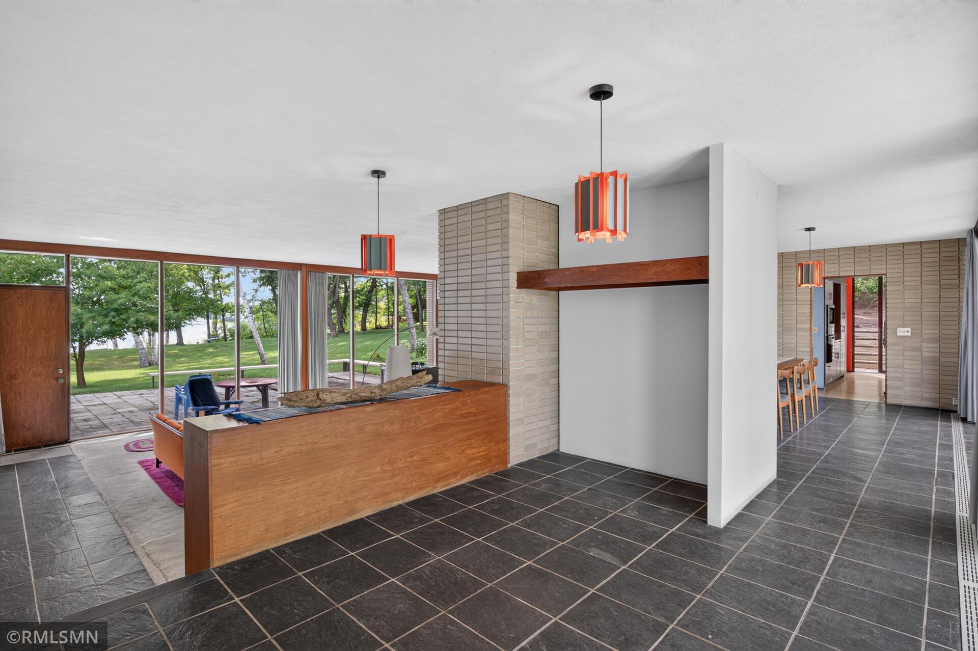 Mid-century living room with a long wood-sided fireplace and windows overlooking a lawn and water.