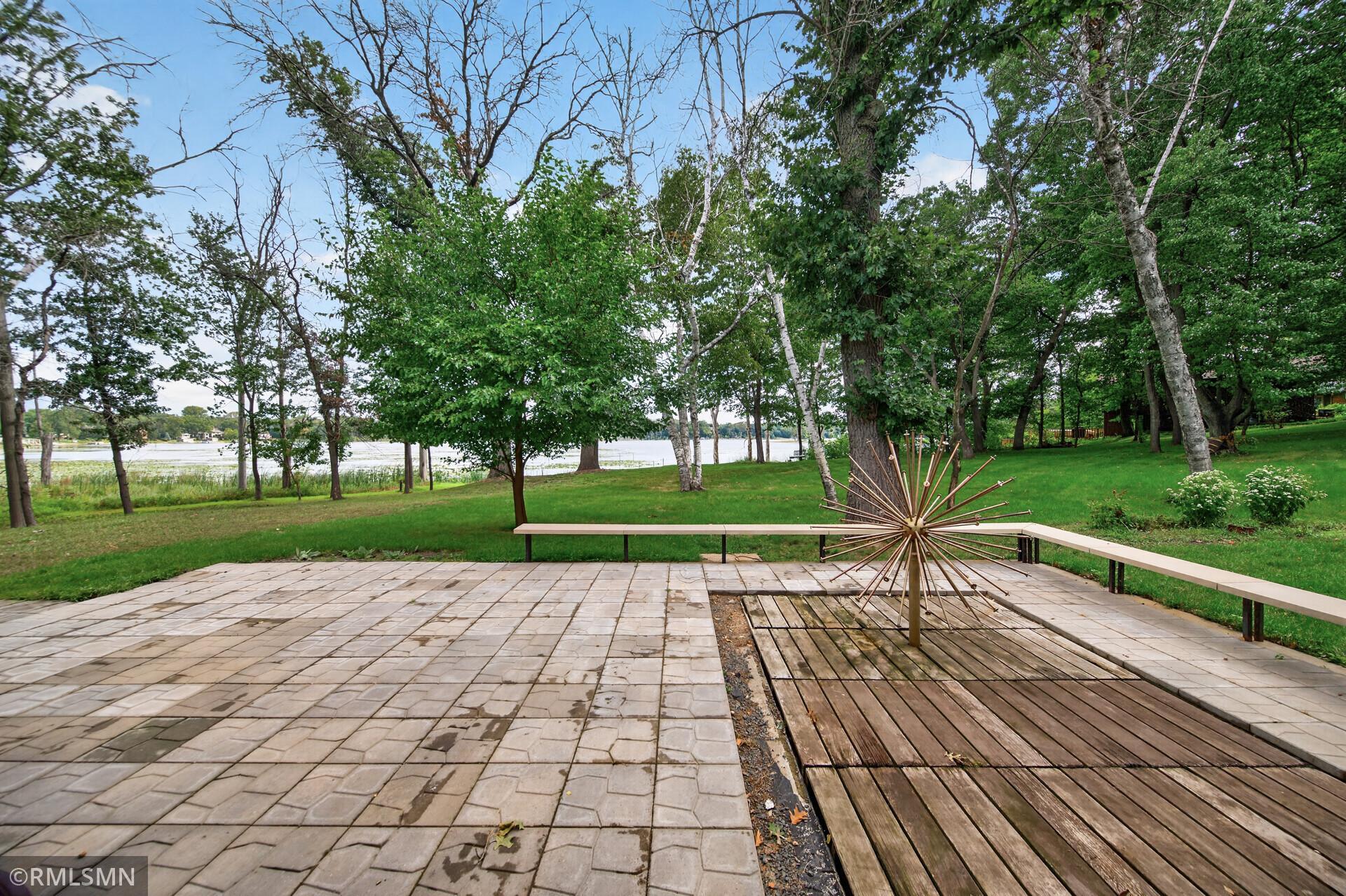 Lakeside backyard patio with a wooden deck, sunburst sculpture, mature trees, and water views.