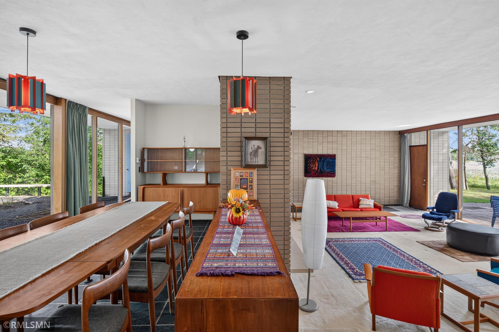 Open-plan dining and living room with colorful pendant lights, a brick column, and large windows overlooking trees and water.