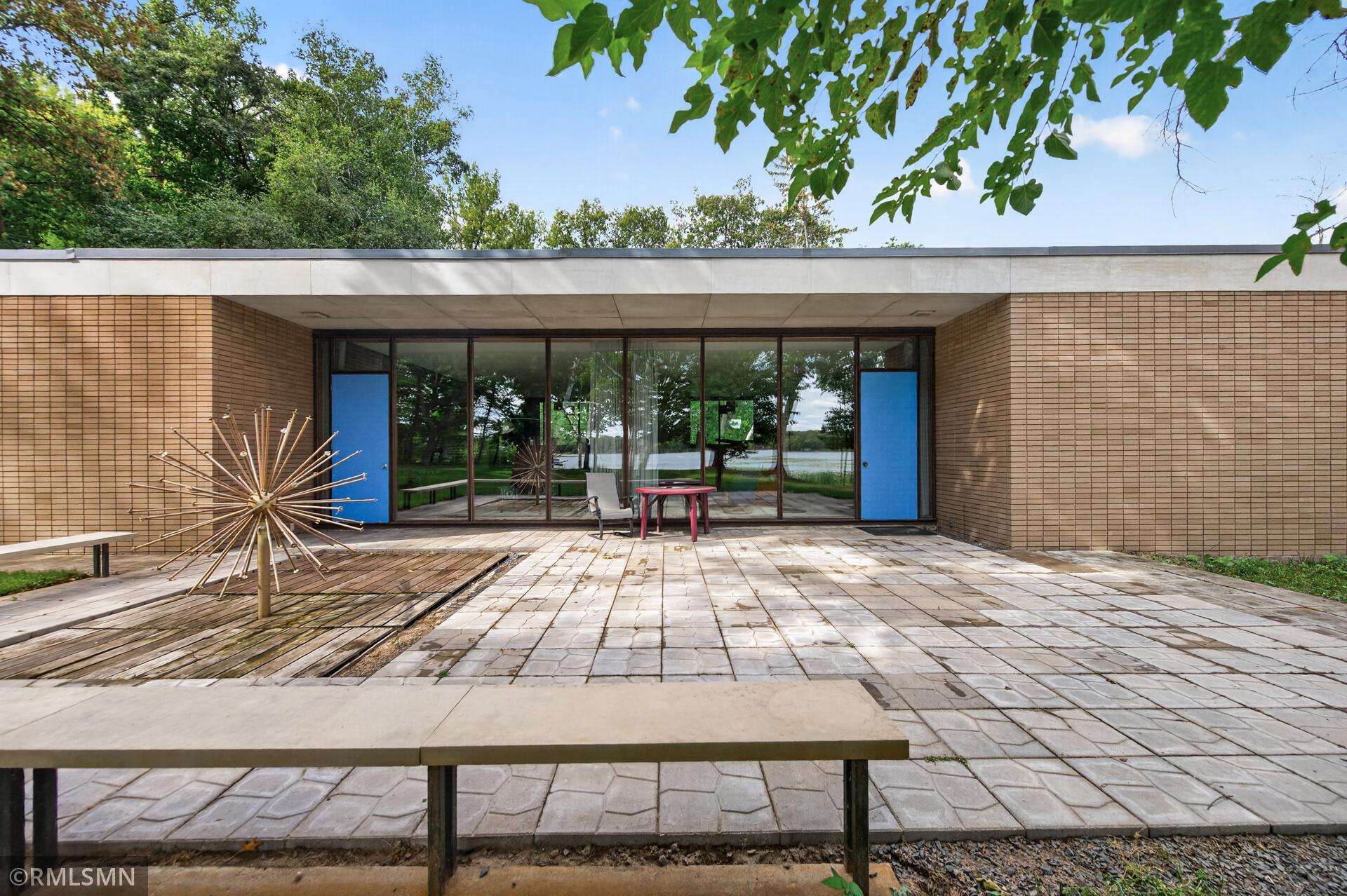 Modern brick home with a glass facade opening onto a paved patio overlooking a lake.