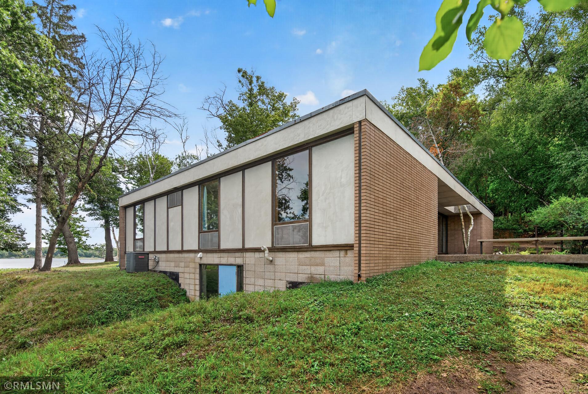 Modernist house with broad windows and brick siding beside a lake, surrounded by mature trees.