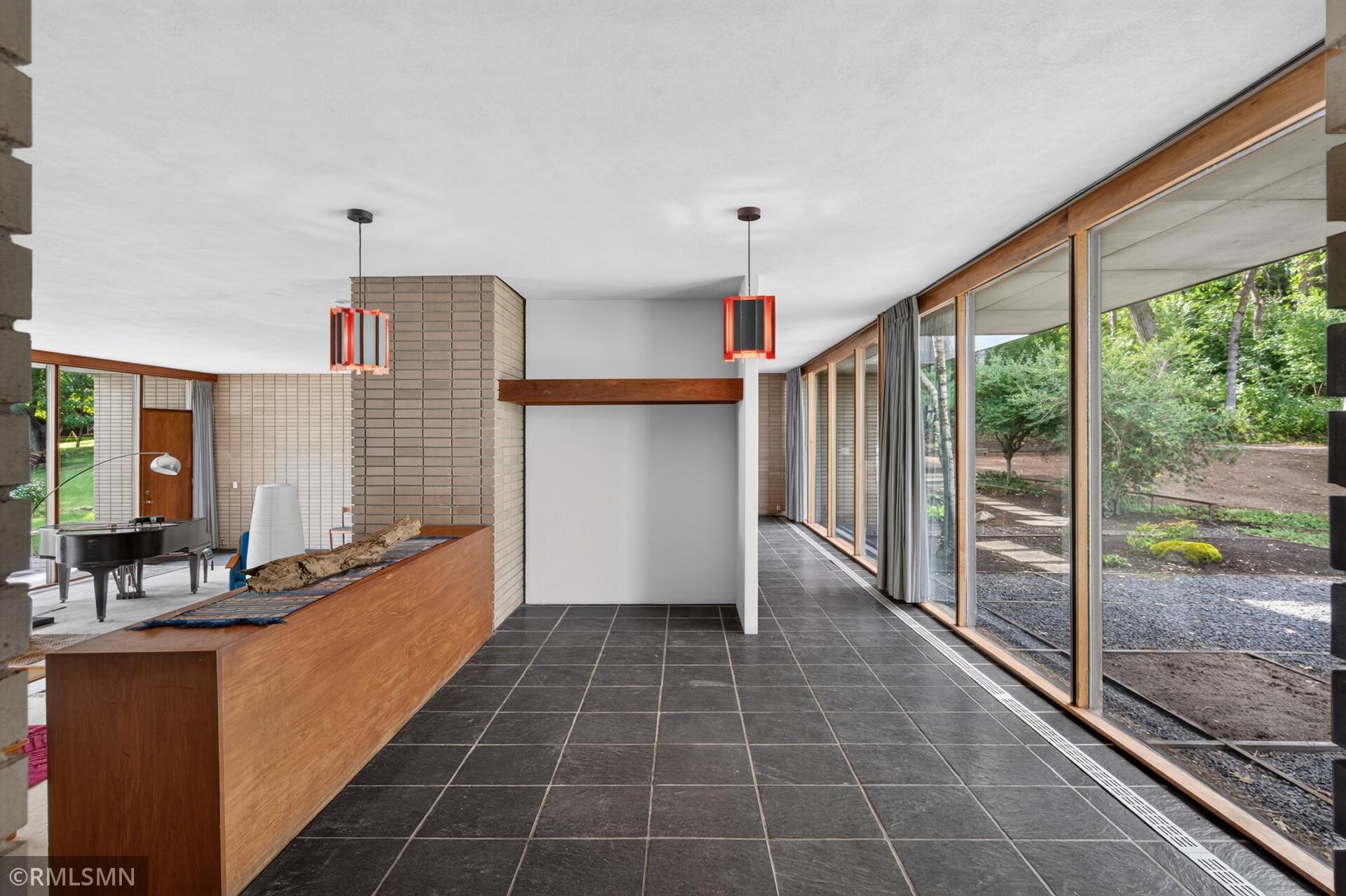 Mid-century living room with a brick fireplace, wood hearth, grand piano, and floor-to-ceiling windows.
