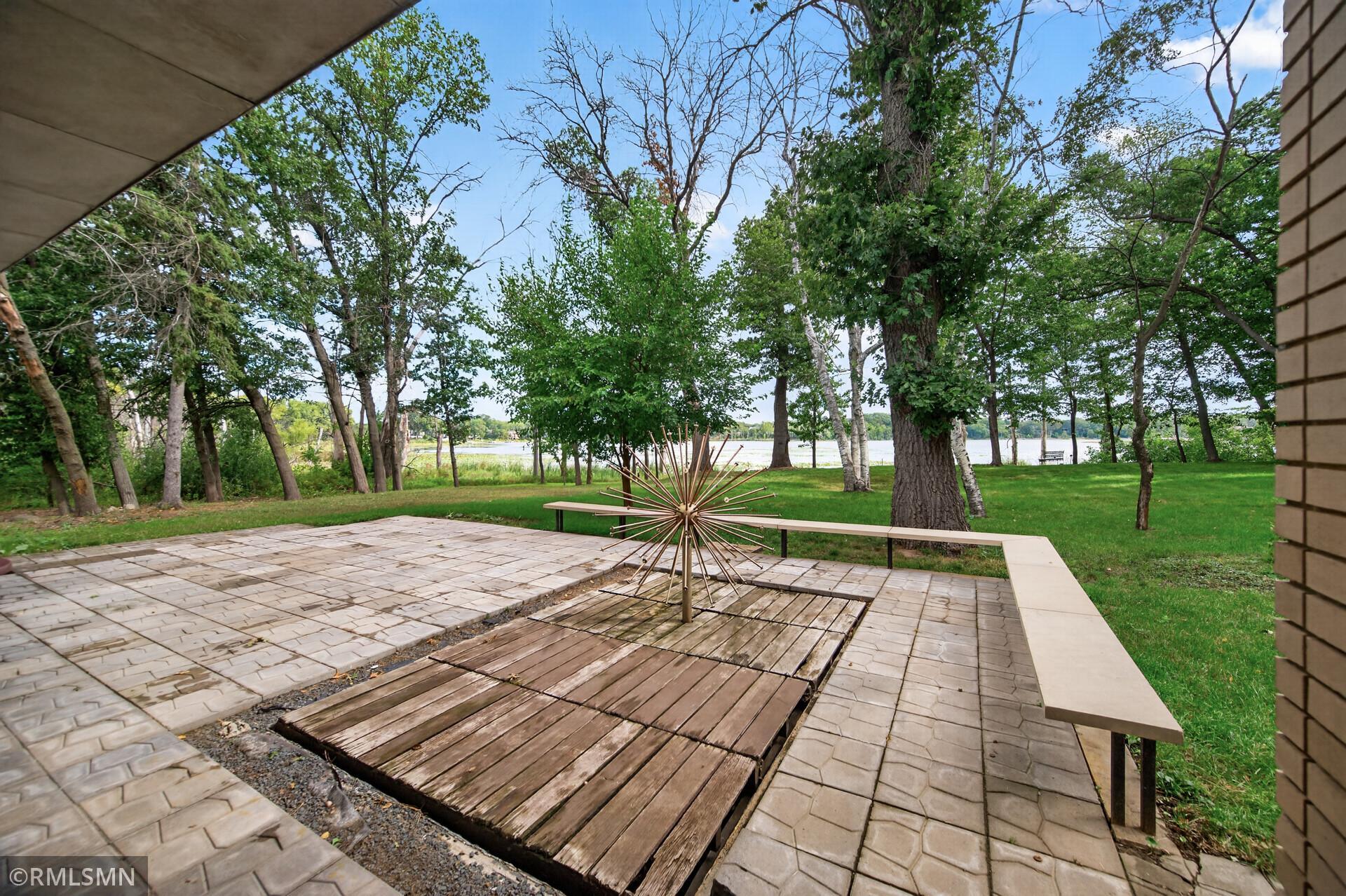 Lakeside patio with stone pavers, wooden deck, benches, and a metal sunburst sculpture beneath mature trees.