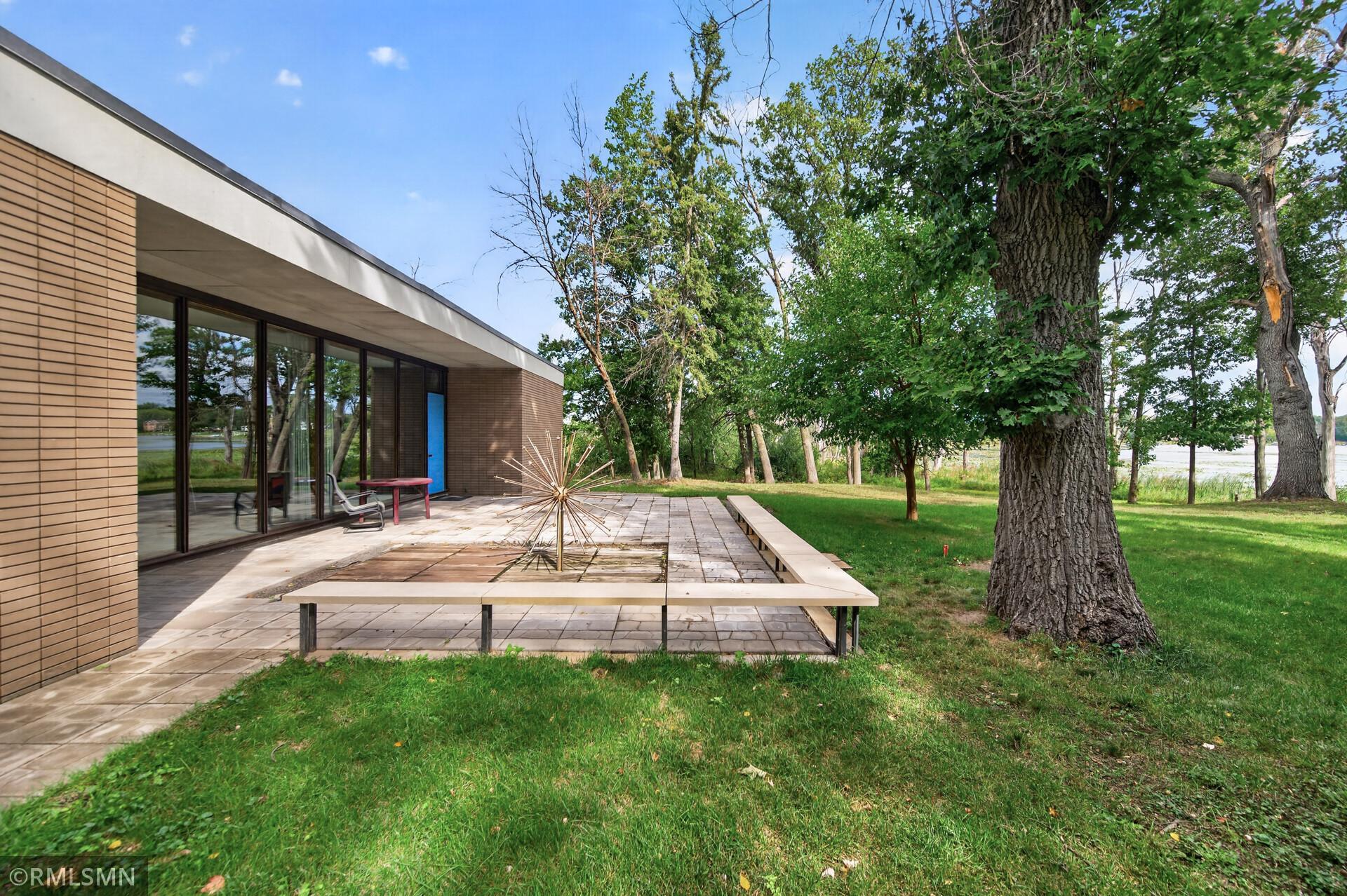 Modern brick home with a paved patio, long bench, and mature trees beside a grassy yard.