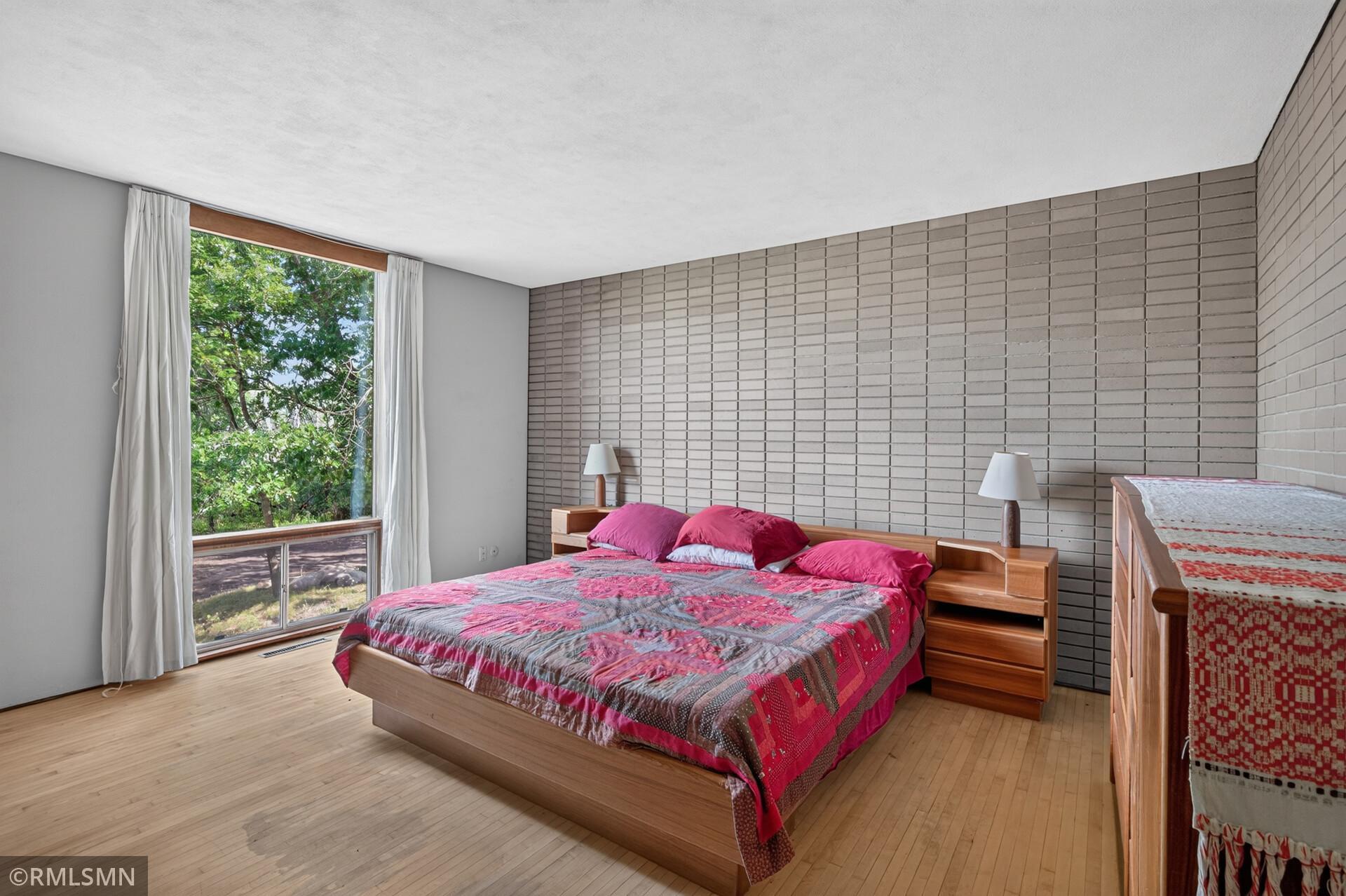 Bedroom with a pink quilted bed, brick accent wall, and large window overlooking leafy trees.