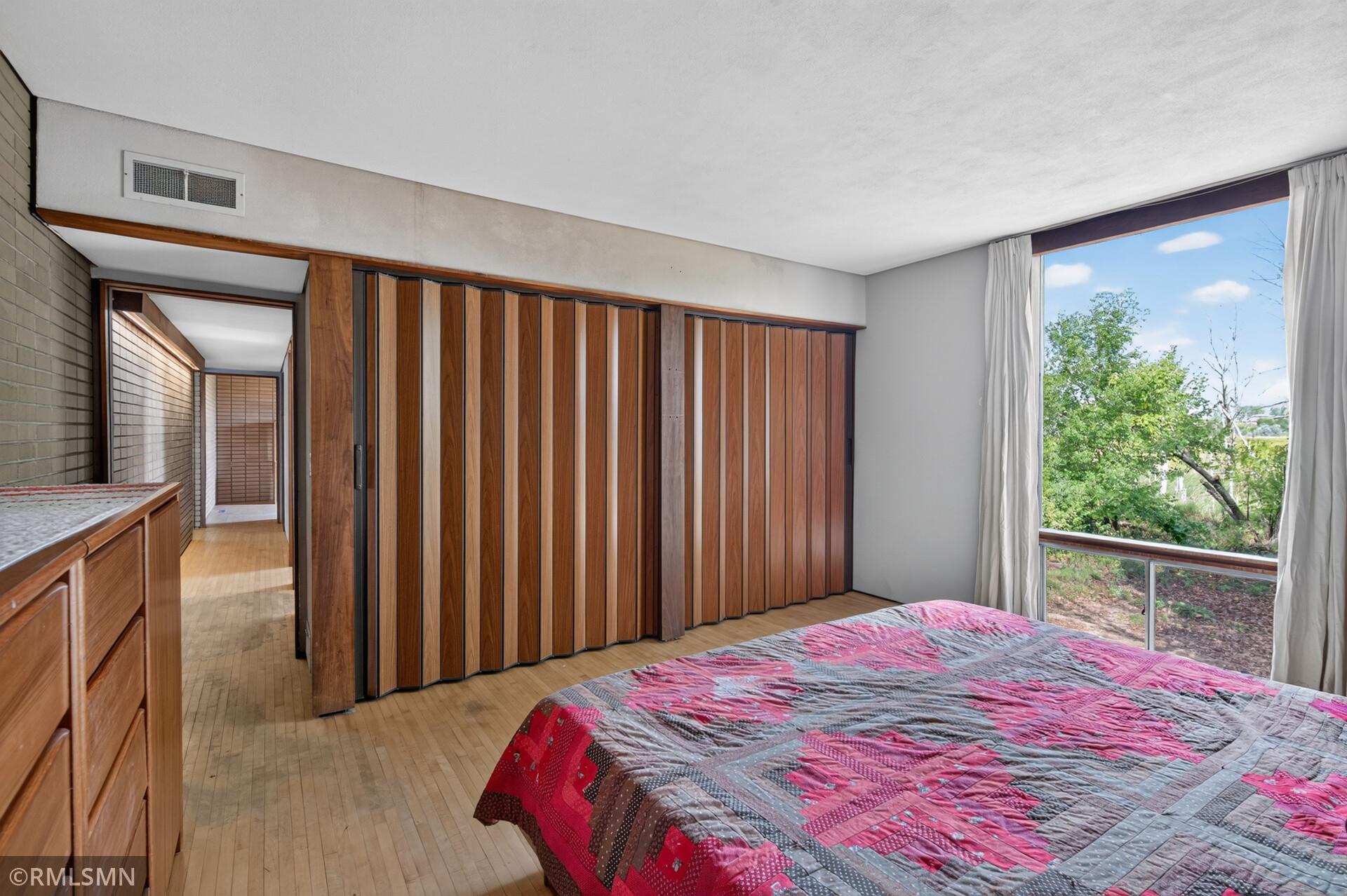 Bedroom with wood folding closet doors, light wood floors, and a large window overlooking trees.