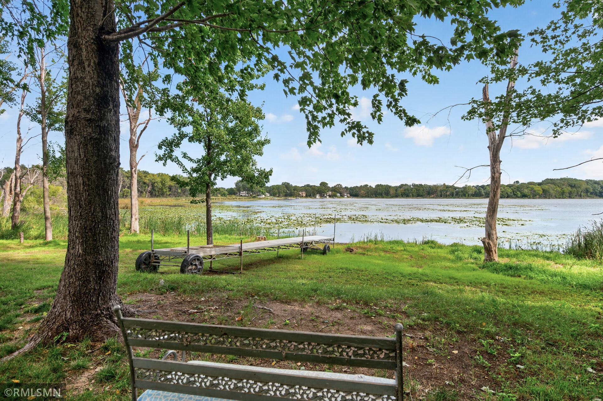 Shaded lakeside lawn with a metal bench and dock beside a lily-pad-covered lake.