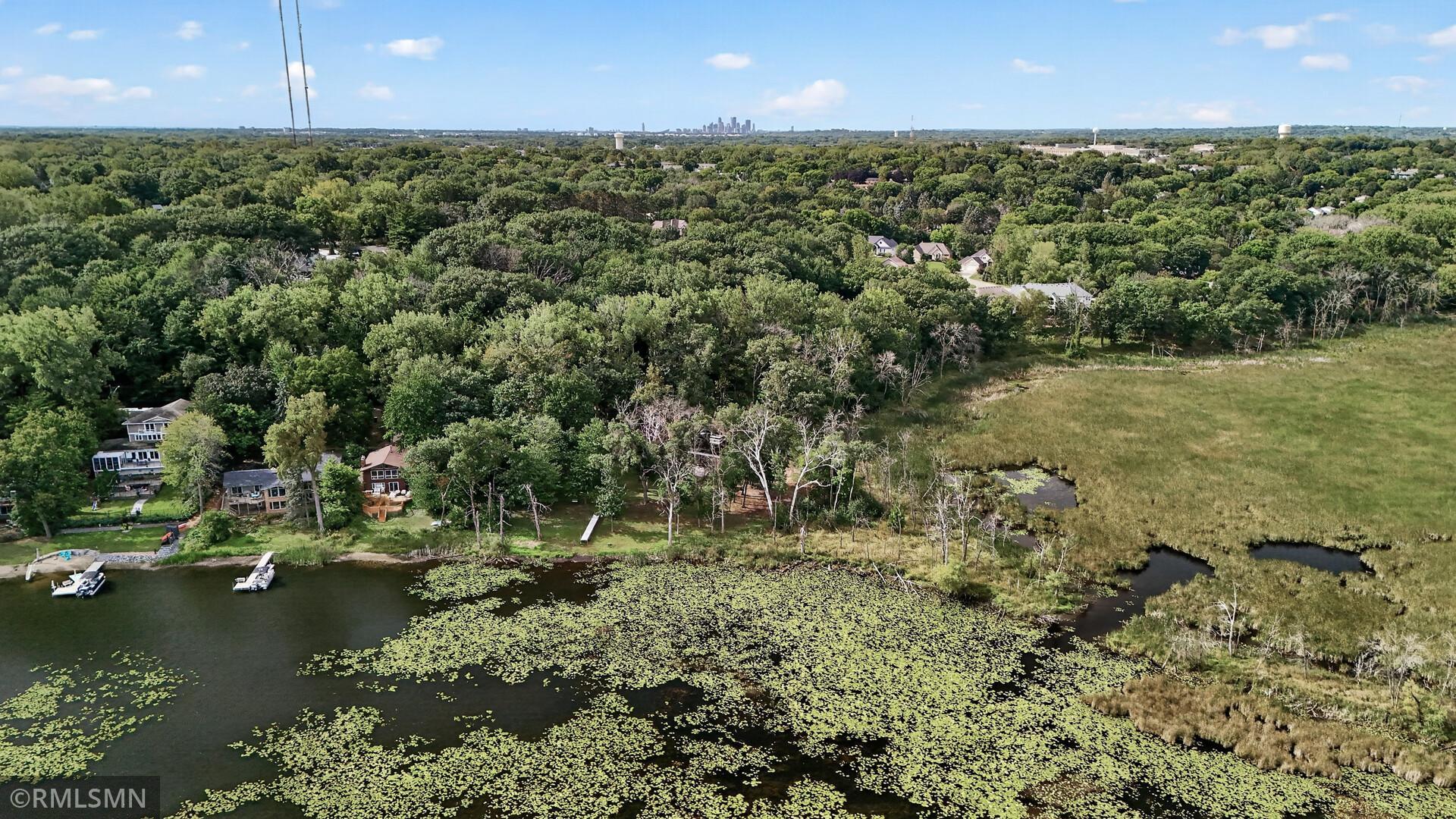 Aerial view of a lake, lily-pad-covered water, wooded shoreline, wetlands, and distant city skyline.