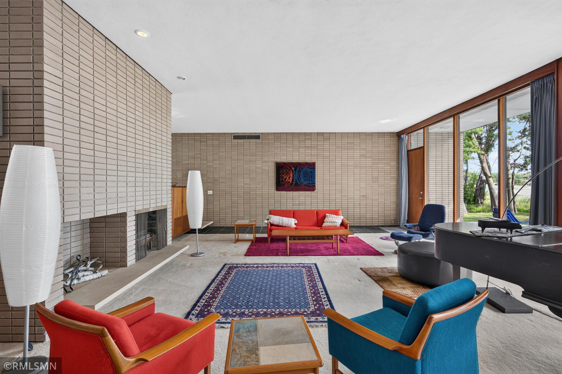 Living room with a brick fireplace, colorful seating, and tall windows overlooking trees.