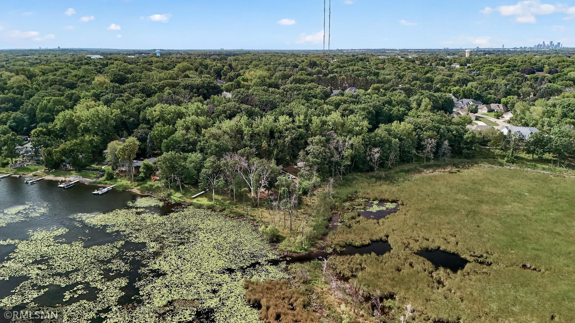 Aerial view of a lily-pad-covered lake, marshland, wooded shoreline, and distant city skyline.