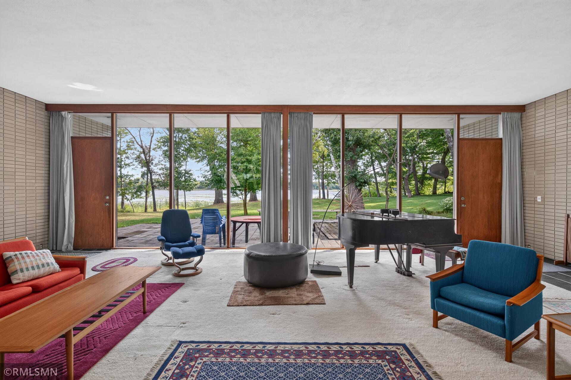 Living room with a grand piano and colorful seating overlooking a leafy lawn and water.