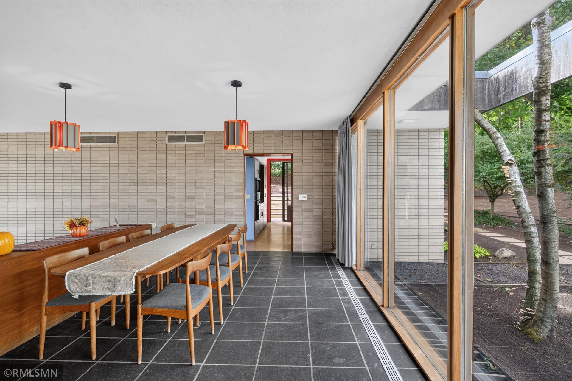Dining room with a long wooden table, orange pendant lights, and floor-to-ceiling windows overlooking trees.