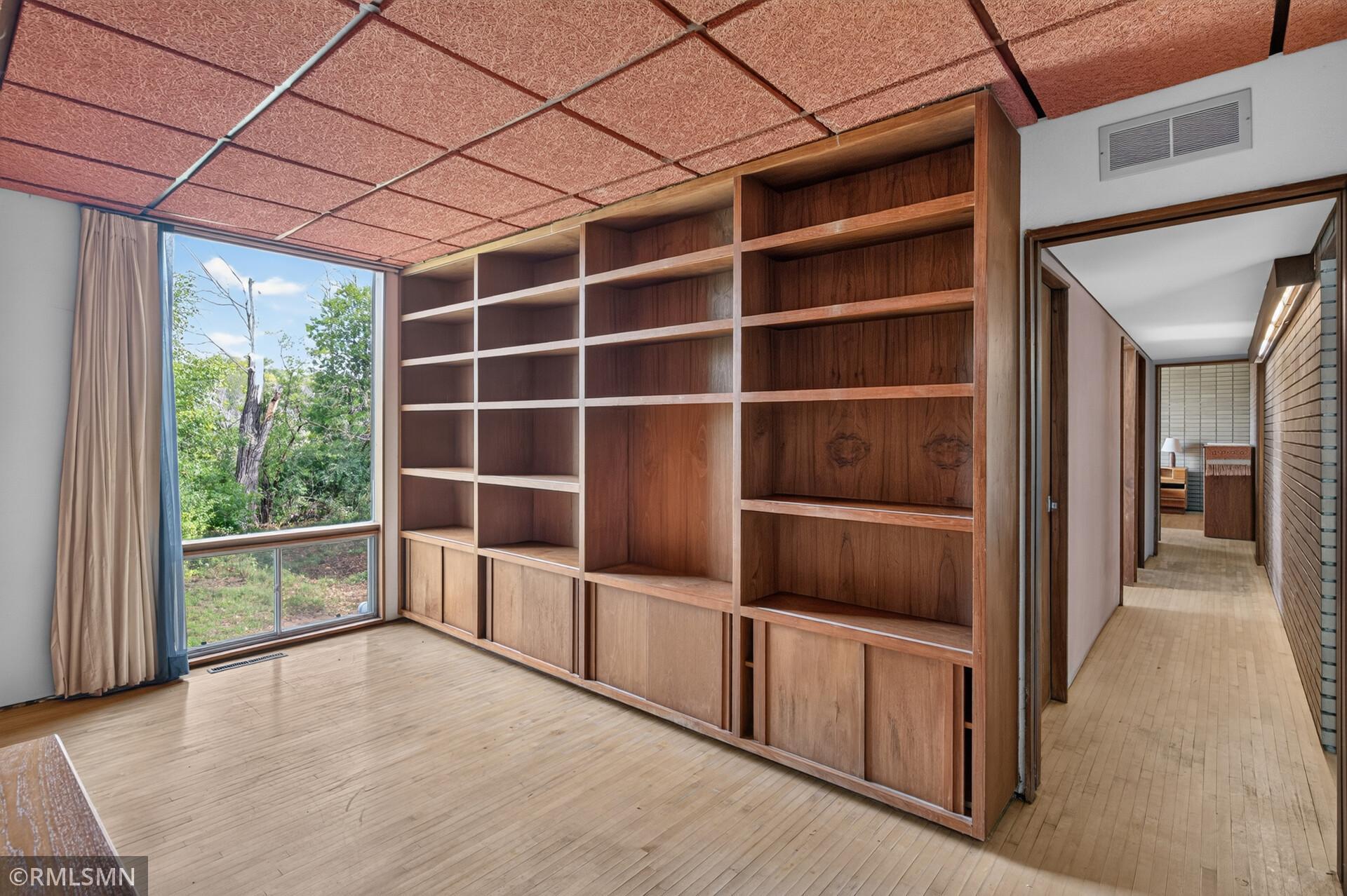 Empty room with built-in wood shelving, light wood floors, and a large window overlooking greenery.