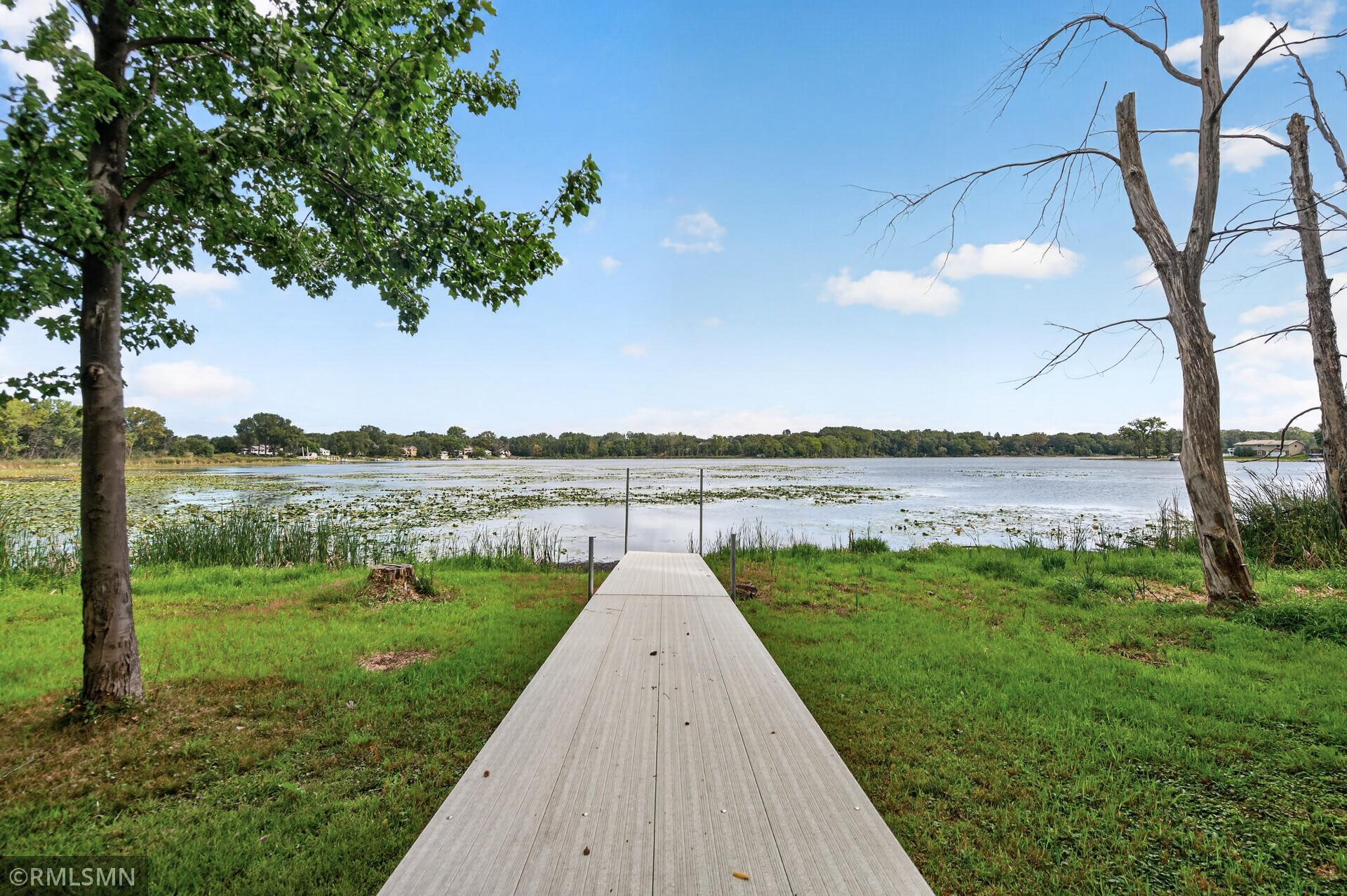 Lakefront dock extending across a grassy shoreline toward a lake framed by trees.