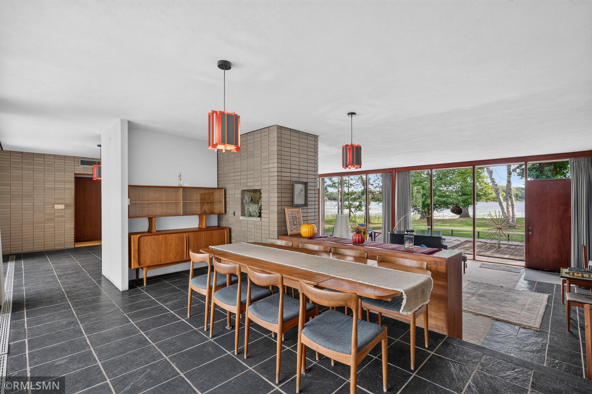 Dining room with mid-century wood furniture, a brick fireplace, and large windows overlooking the water.