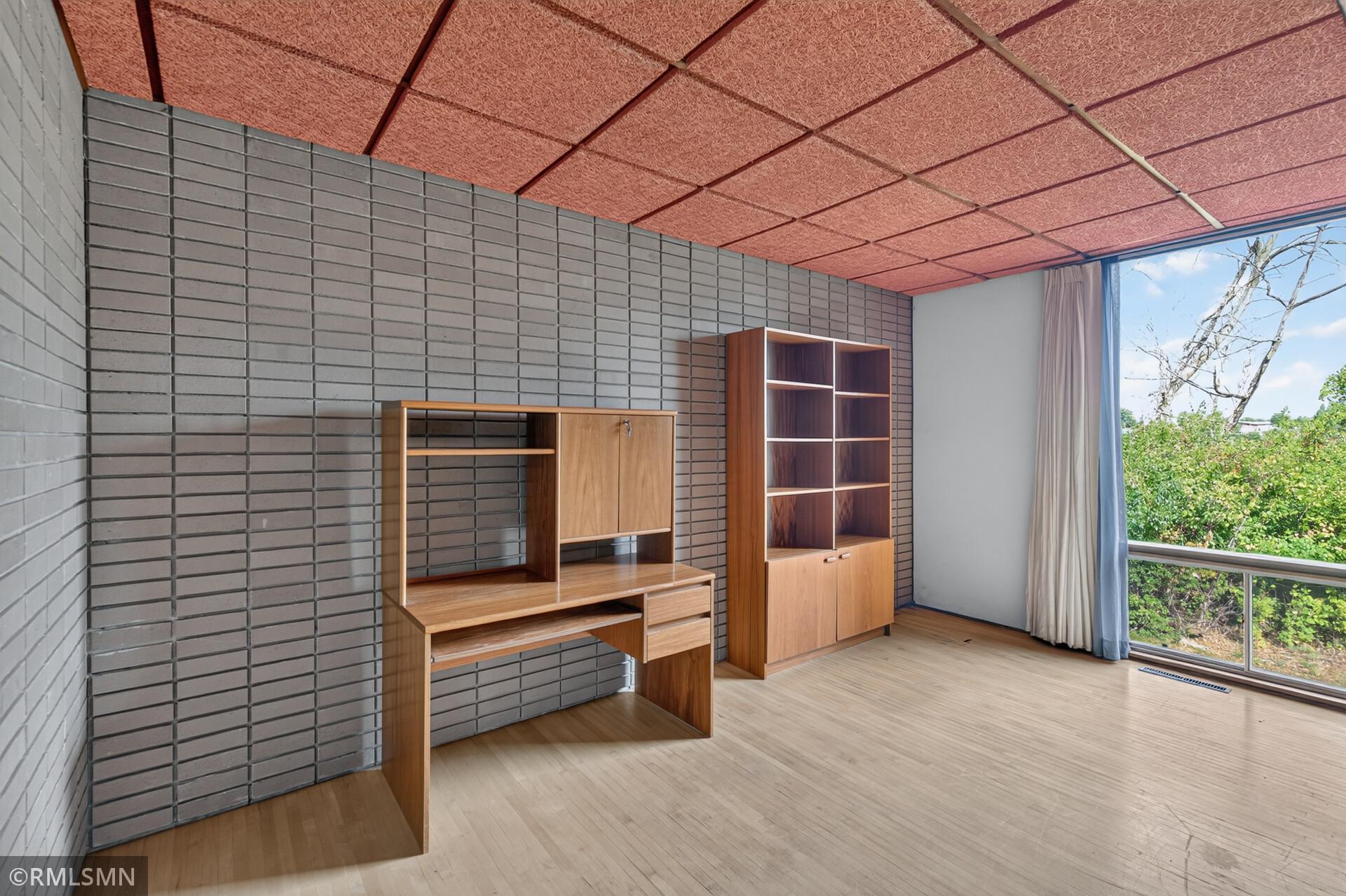 Room with a built-in wooden desk and shelving, brick walls, and a wide window.