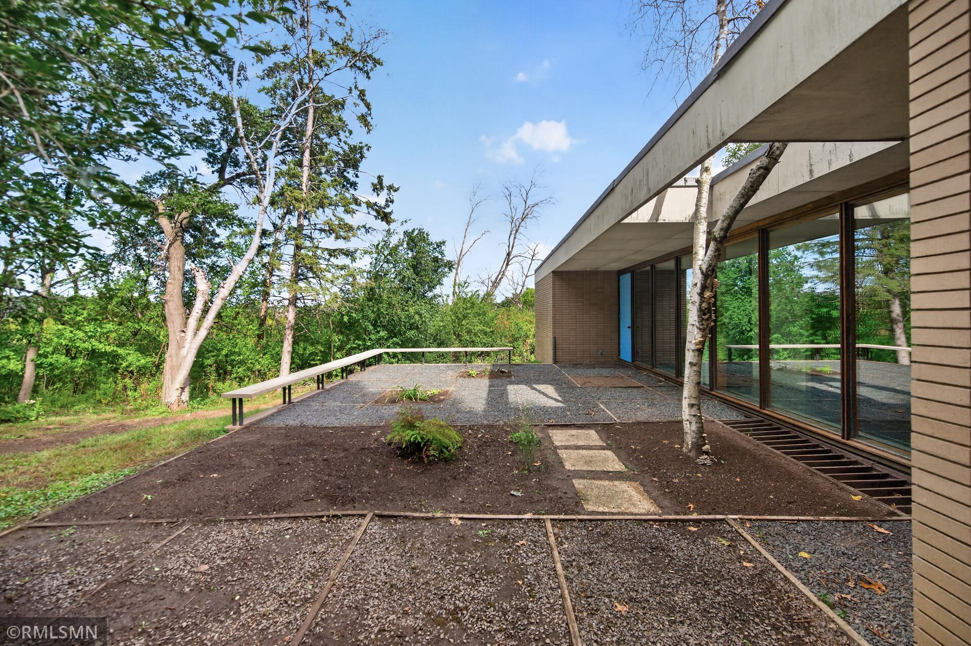 Modern home exterior with a broad gravel terrace, low railing, and large windows facing wooded surroundings.