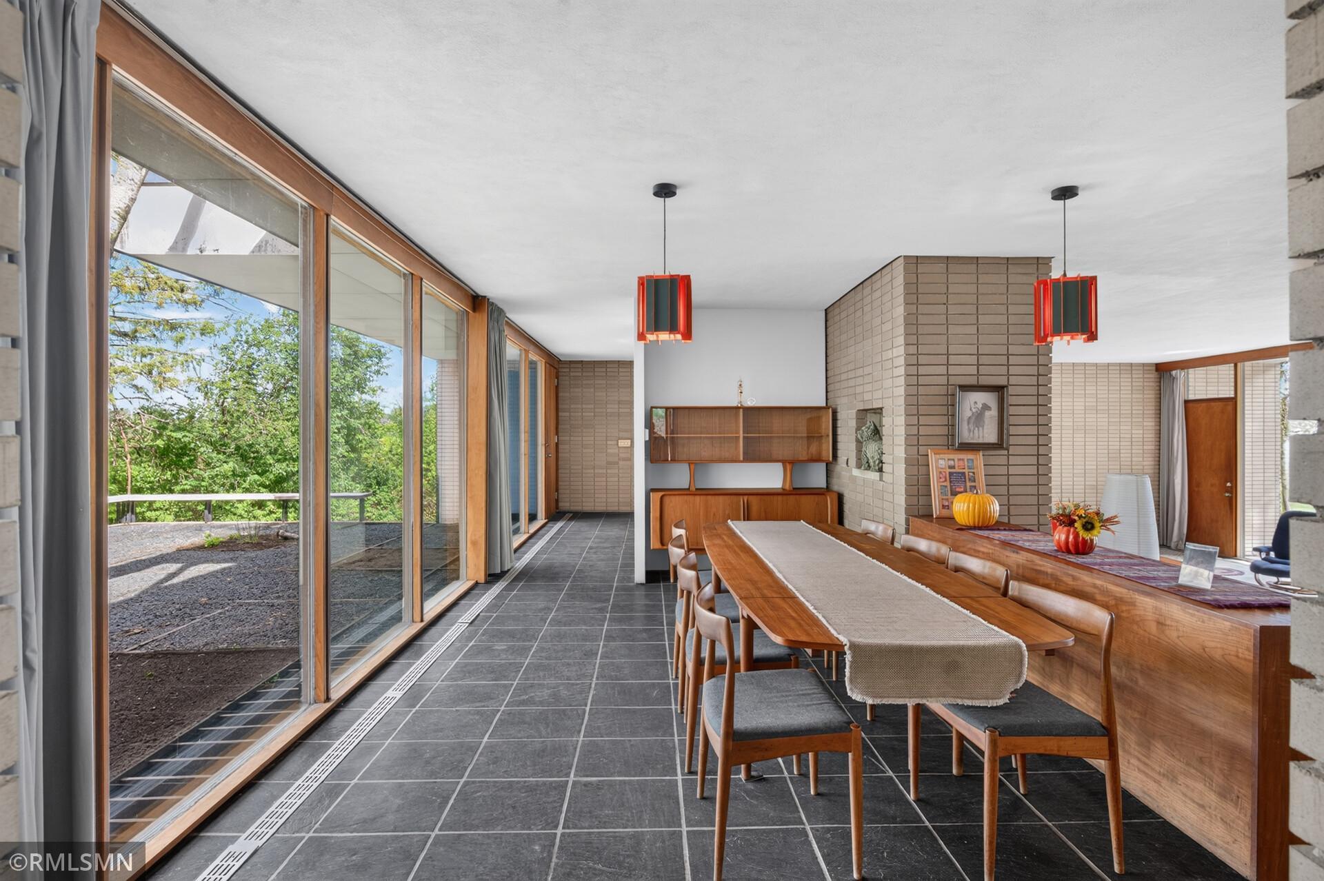 Dining room with a long wooden table, red pendant lights, brick walls, and expansive windows.