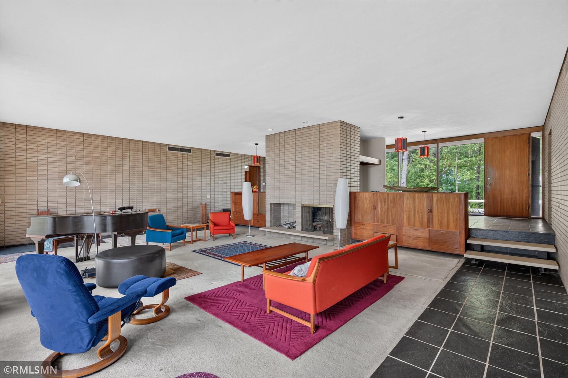 Living room with brick walls, a fireplace, colorful midcentury furniture, and a grand piano.