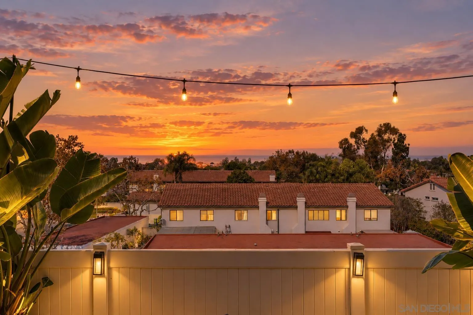 Ocean sunset viewed over tile-roof homes, framed by tropical foliage and glowing string lights.