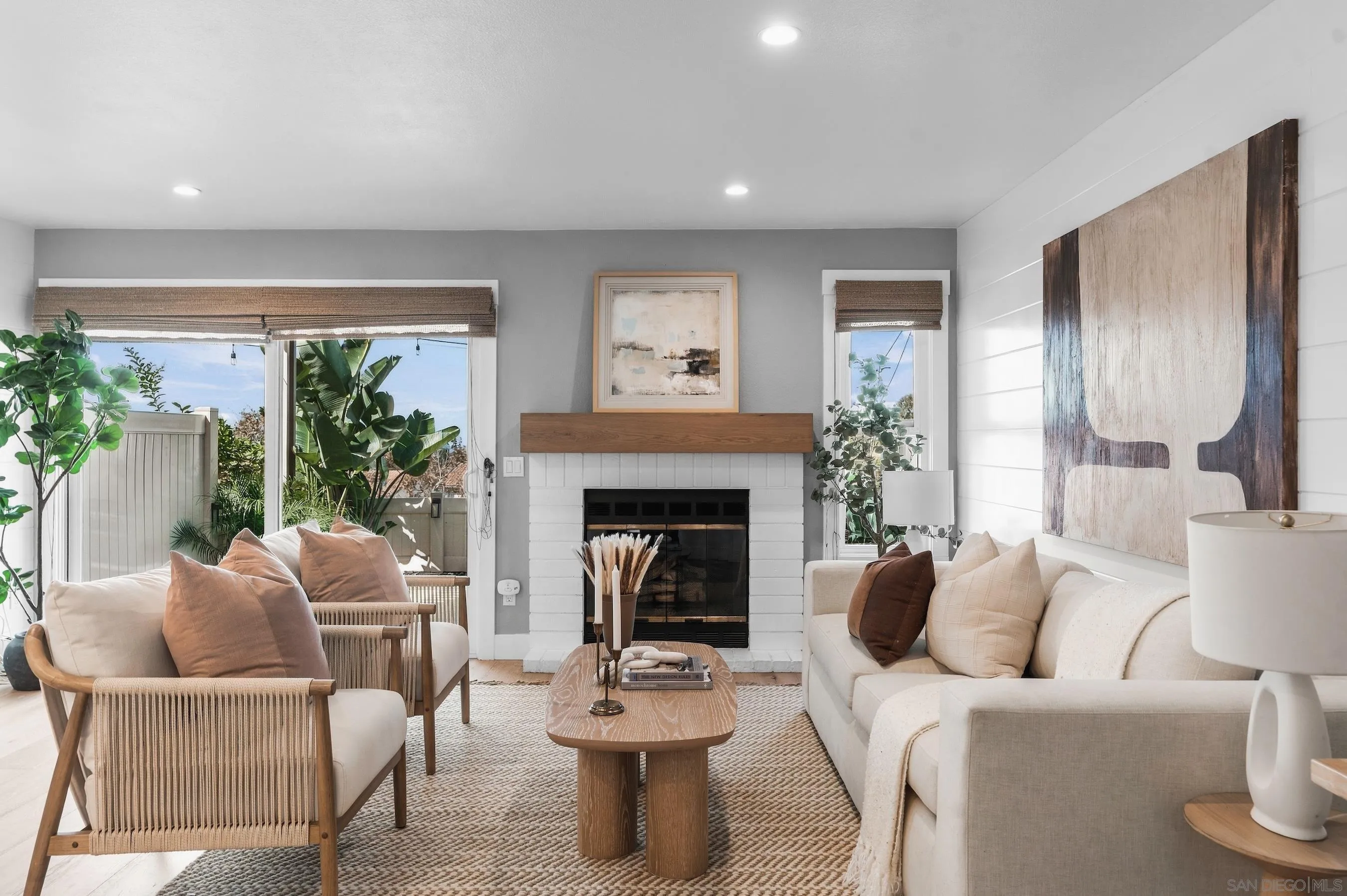 Staged living room with a white fireplace, cream sofas, woven chairs, and windows overlooking a leafy patio.