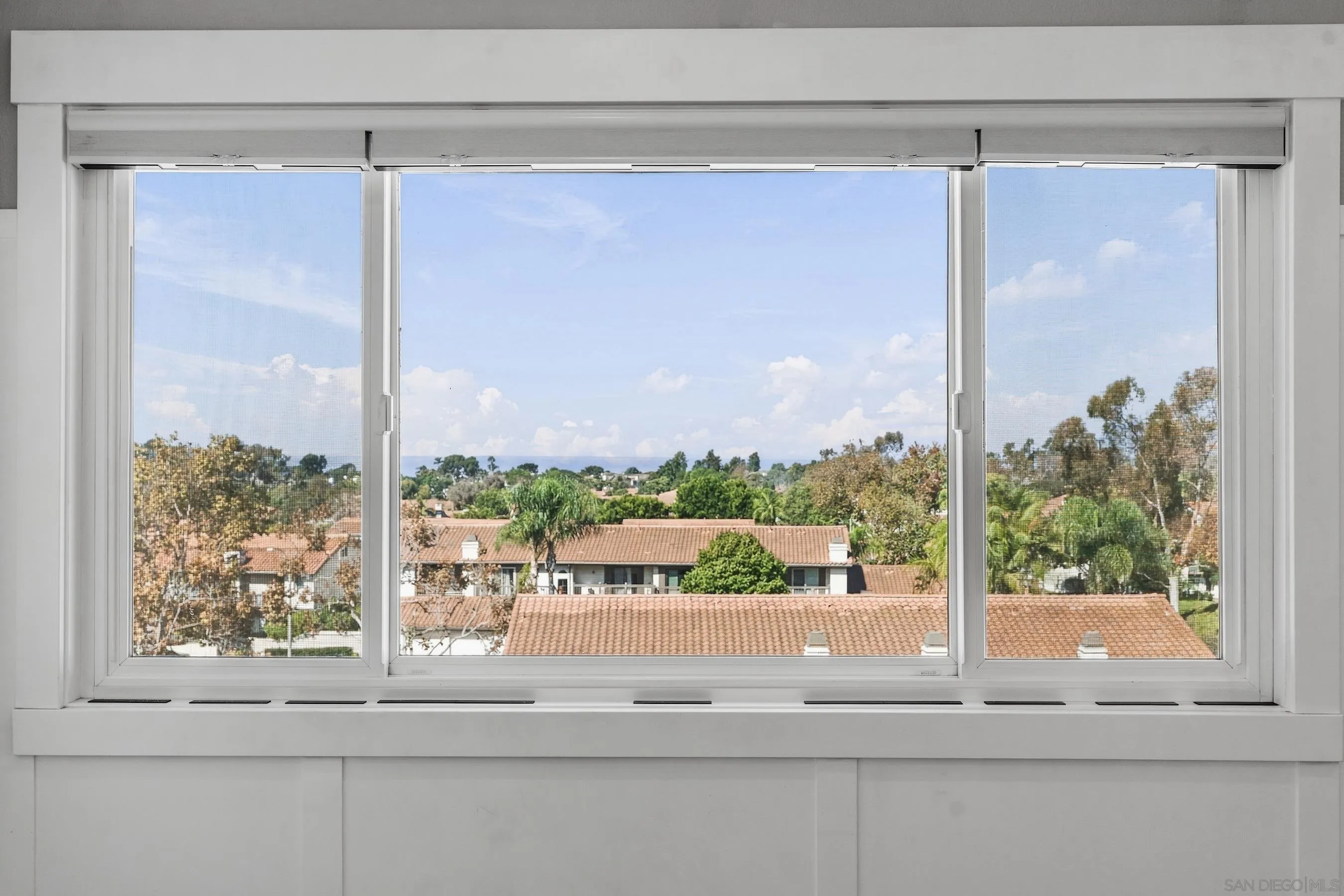 Wide window view over tile-roof homes and leafy trees beneath a bright blue sky.