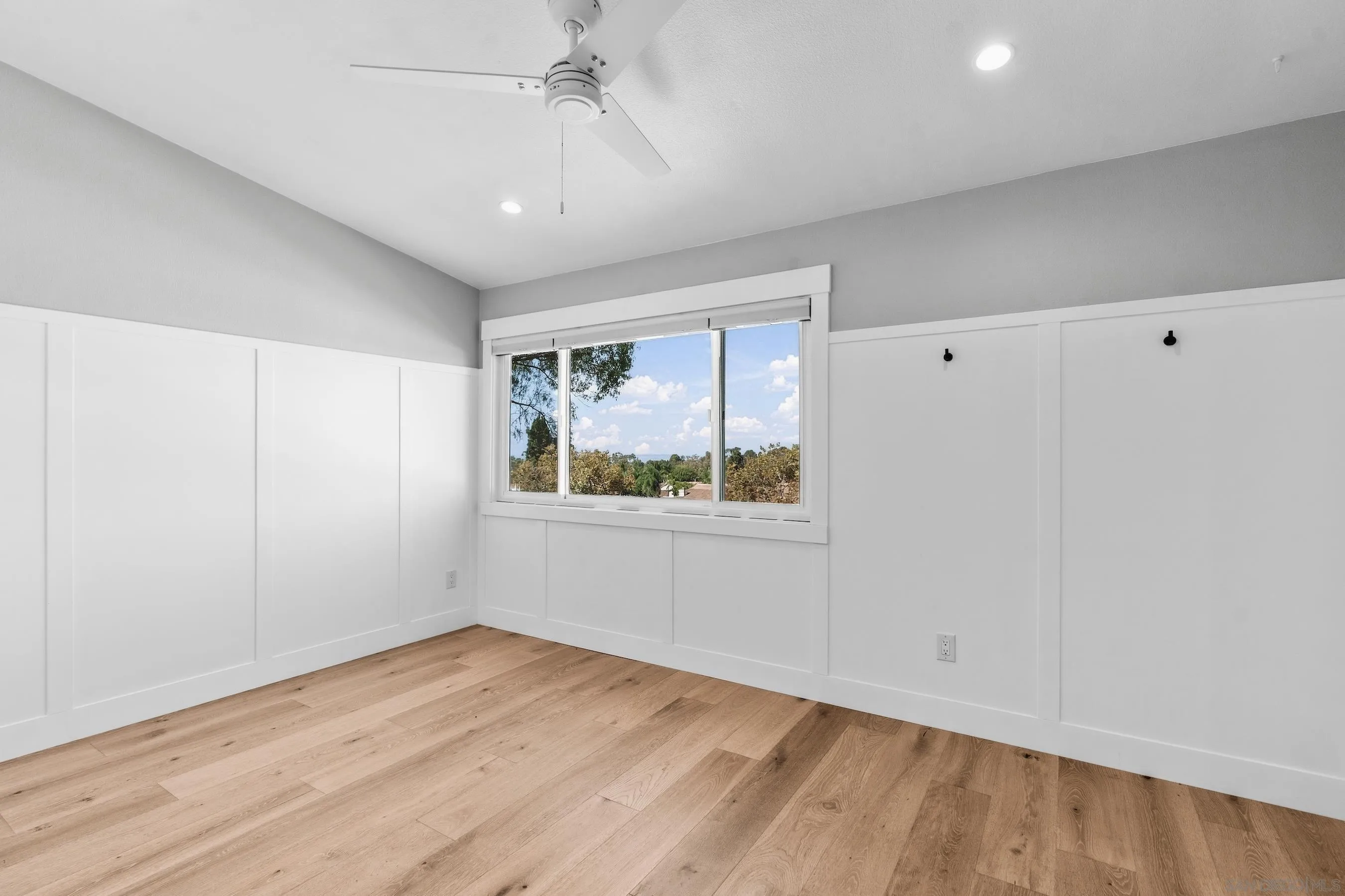 Empty room with light wood floors, white paneled walls, a ceiling fan, and a wide window overlooking trees.