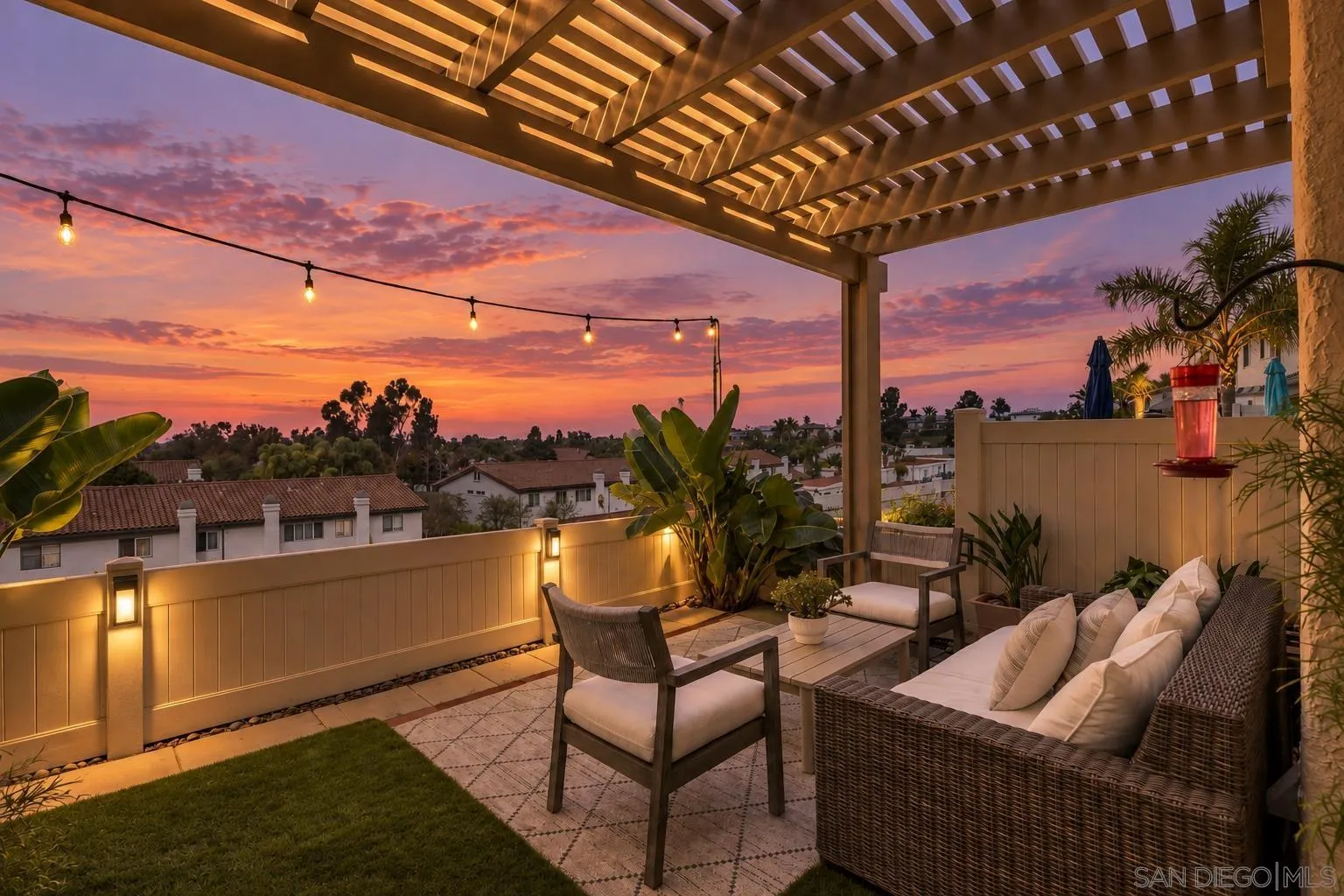 Furnished pergola patio with string lights and a colorful sunset over nearby homes.