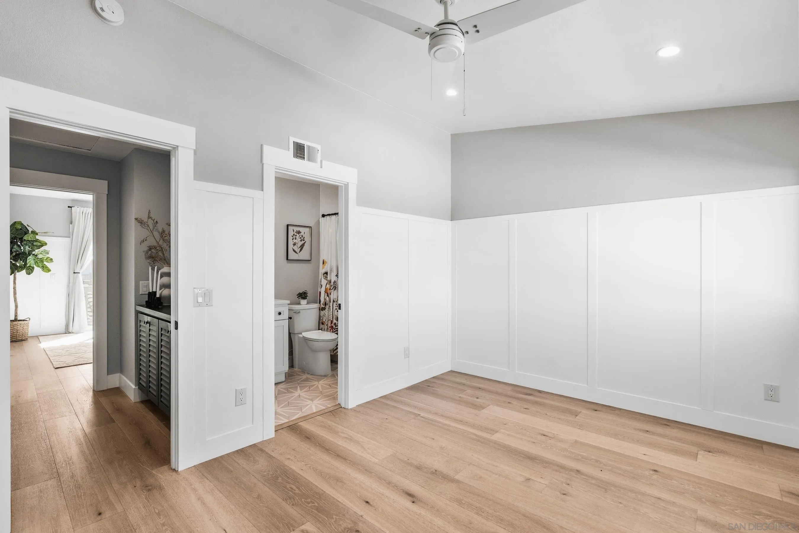 Empty bedroom with light wood floors, white paneled walls, and an open doorway to a tiled bathroom.