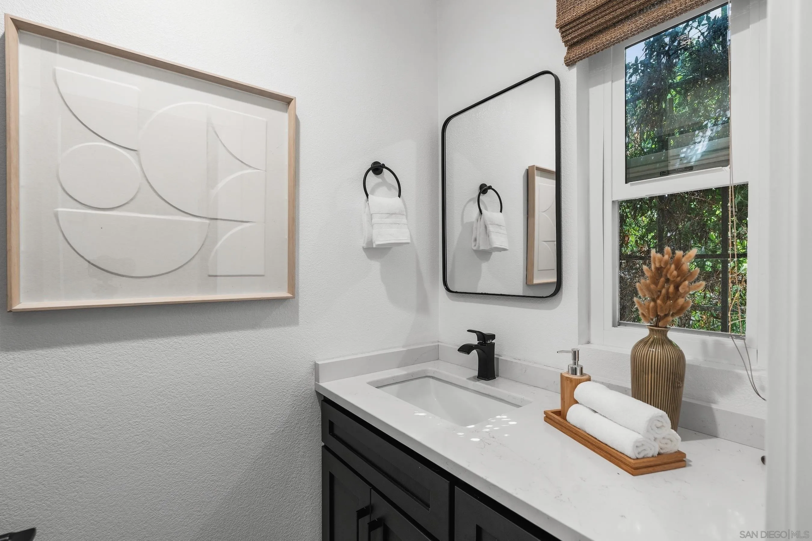 Bathroom vanity with black cabinetry, white countertop, framed art, and a window overlooking greenery.