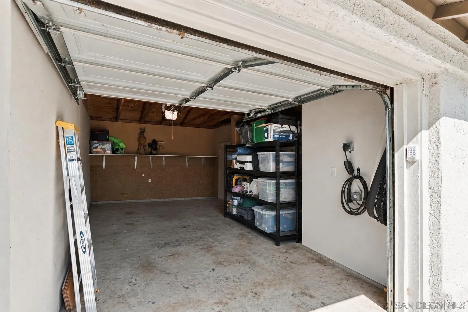 Garage with open overhead door, storage shelving, bins, and a ladder.