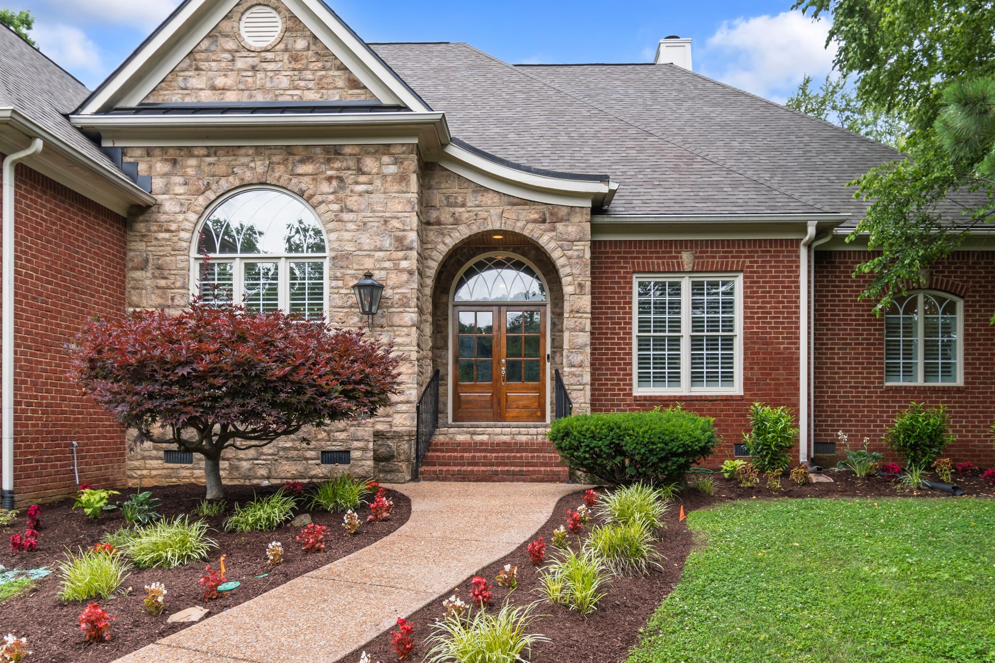 Home exterior with red brick, tan stone, arched windows, and a landscaped curved walkway.