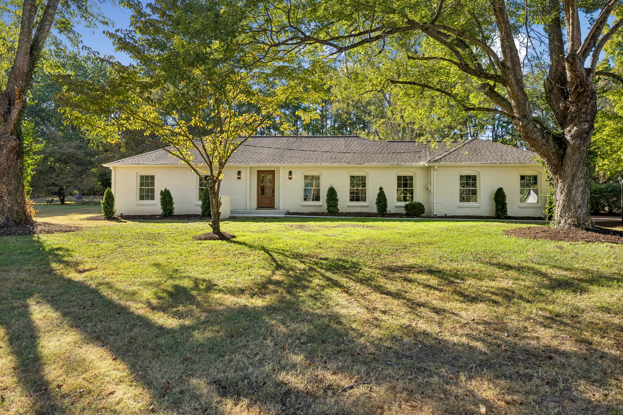 White brick ranch-style home with a central wooden entrance, broad lawn, and mature trees.