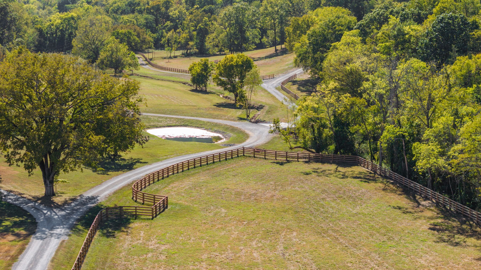 Aerial view of fenced grassy paddocks and a gravel drive beside a small pond and wooded edges.