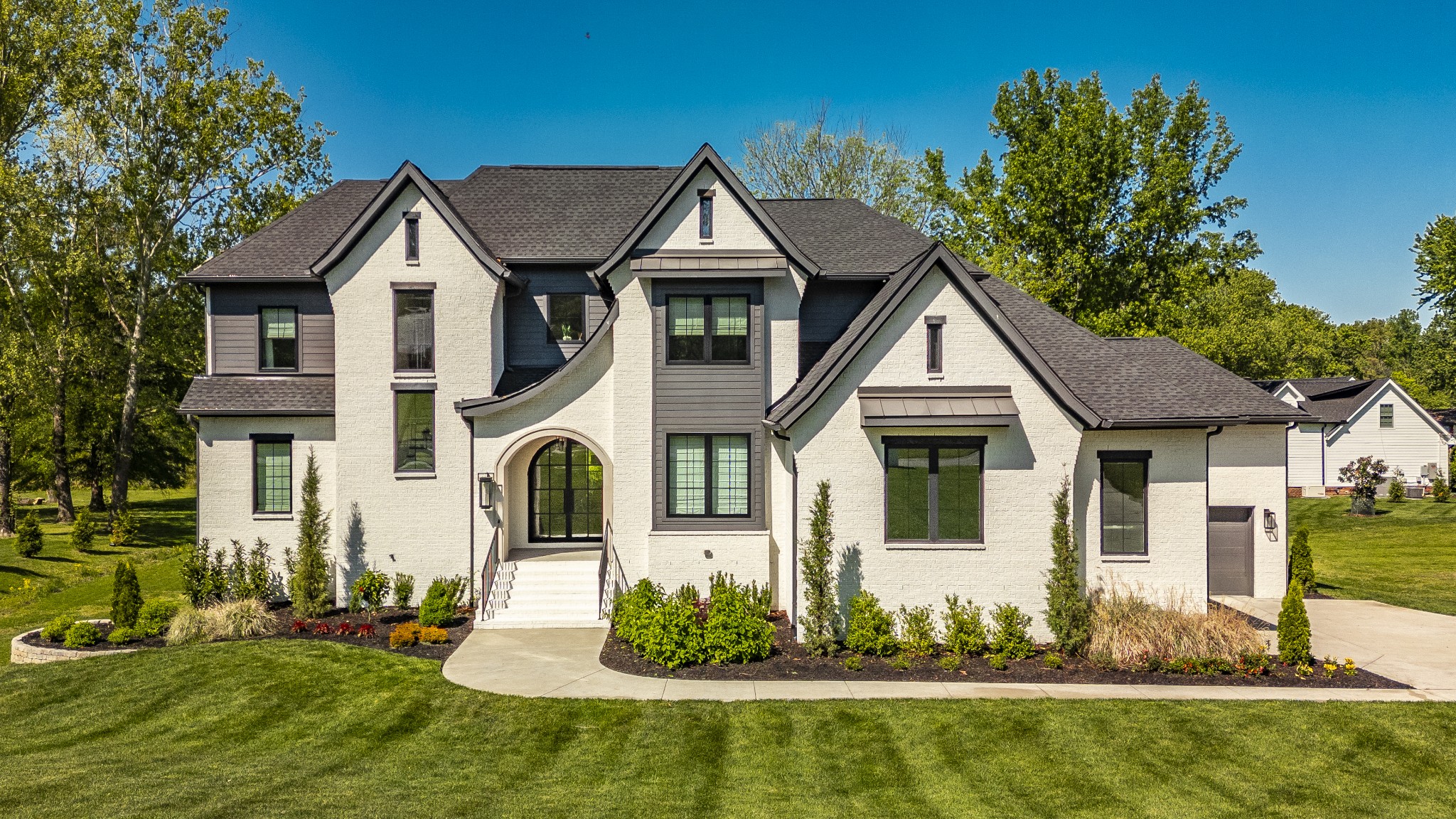 White brick two-story house with an arched entry, dark gabled roof, landscaped lawn, and attached garage.